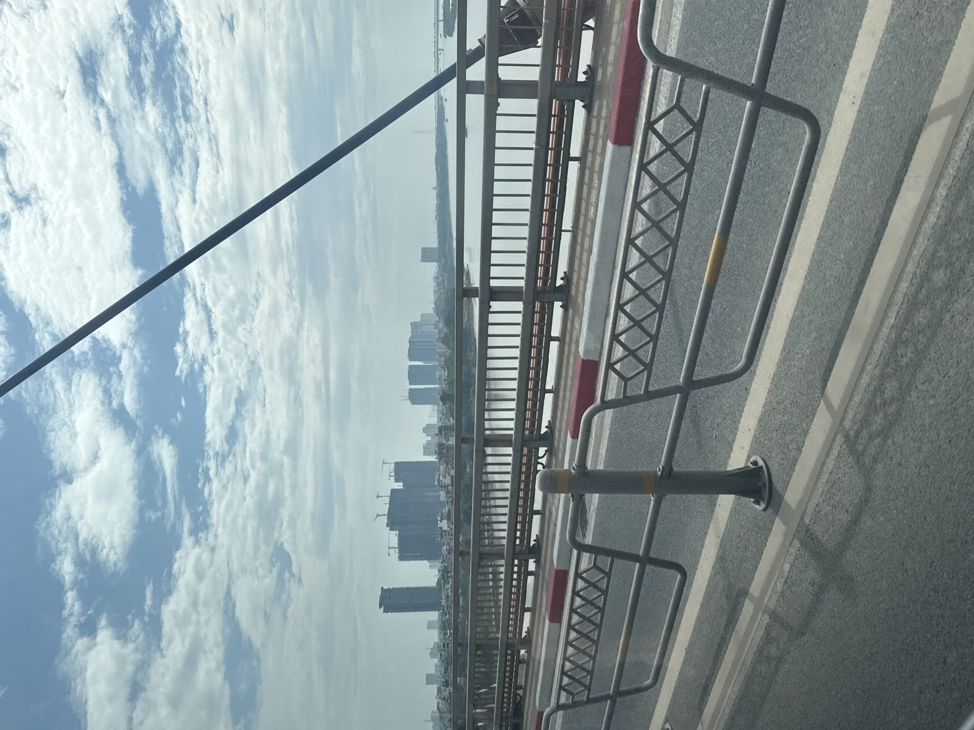 The image shows a view from a bridge in Hà Nội, Vietnam. In the background, there is a hazy cityscape of tall buildings under construction, with several cranes visible. A body of water, likely the Red River, separates the bridge from the city. The bridge itself features gray metal railings with red and white stripes, as well as gray metal safety barriers along the roadway. The road surface is gray asphalt with white lane markings. A thick black cable runs diagonally across the upper left portion of the frame, obscuring the view of the sky, which is mostly bright with scattered clouds.