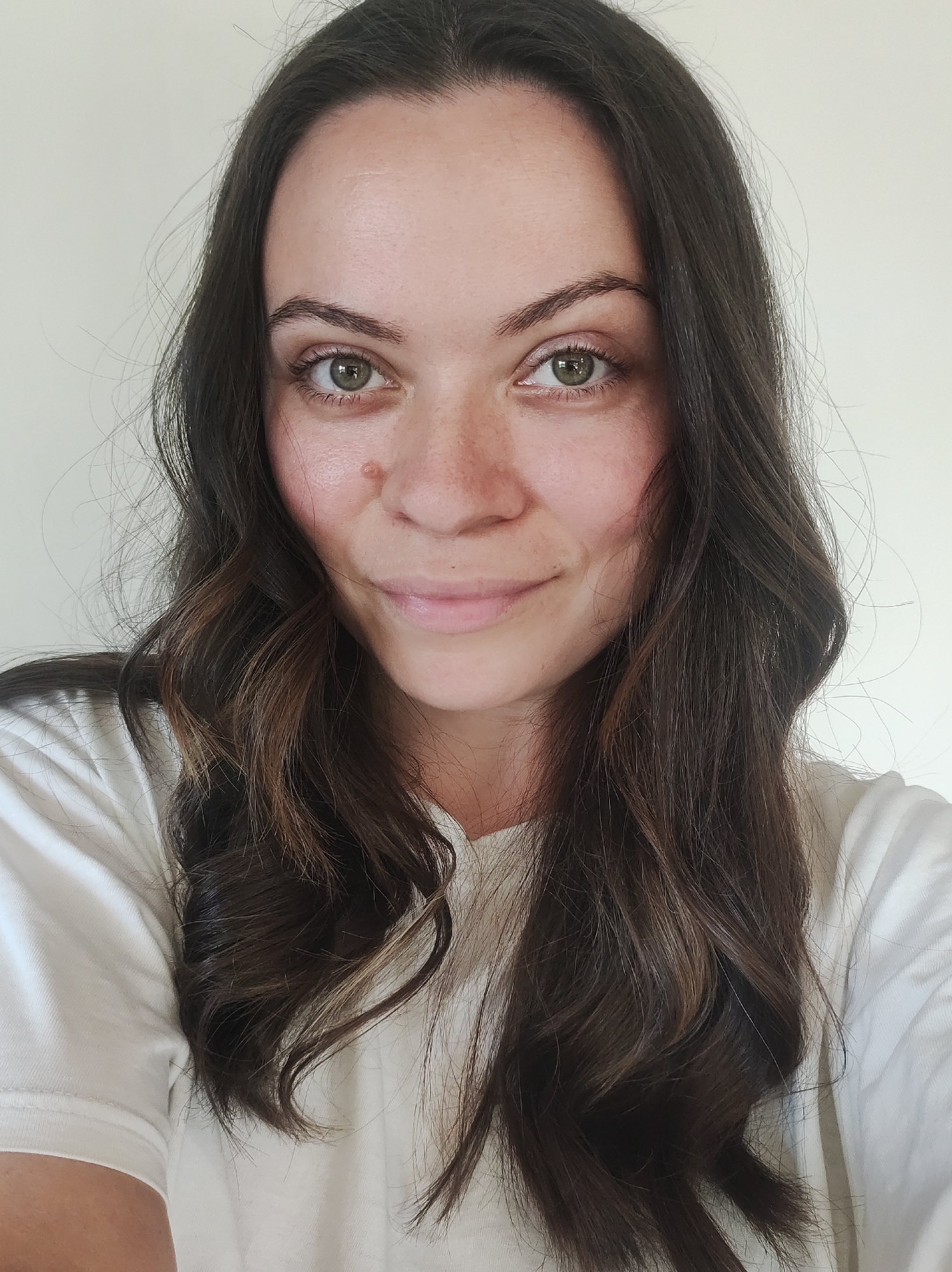 A close-up portrait shows a woman with long, wavy dark brown hair. Her hair has lighter brown highlights. She has light skin, hazel eyes, and a mole on her right cheek. She wears a white t-shirt. The background is a plain, light-colored wall. The location is Unknown, Unknown.
