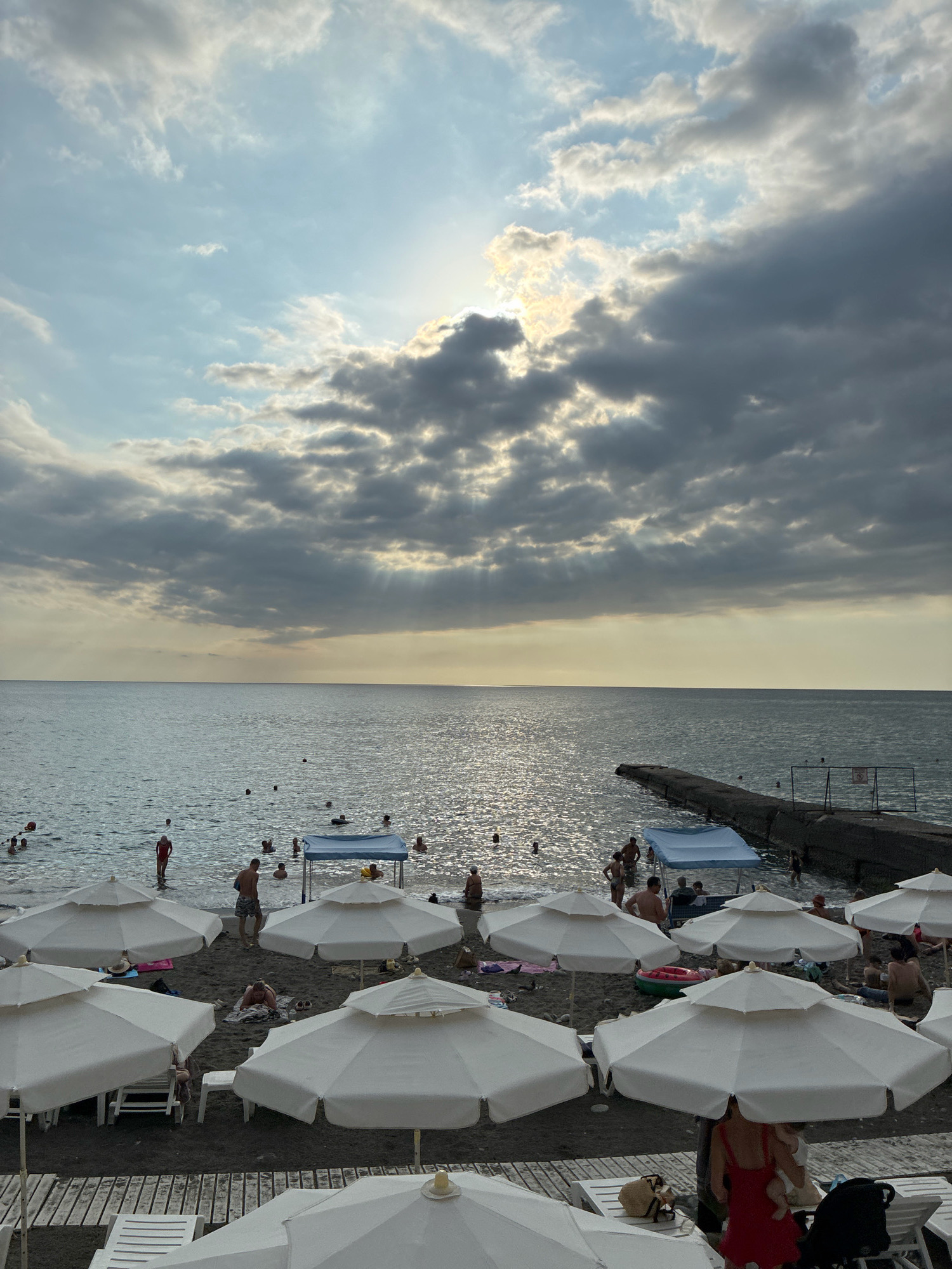 The image depicts a beach scene in Sochi, Russia, on a cloudy day with sunbeams breaking through the overcast sky. The sea is calm, with several people swimming and wading near the shore and a long concrete pier extending into the water. The beach itself is populated with numerous white beach umbrellas, some open and providing shade, while others are closed. Beach chairs and towels are visible on the dark sand. A few individuals are seen relaxing under the umbrellas, while others are walking or standing on the beach. The overall atmosphere suggests a leisure activity on a coastal area.
