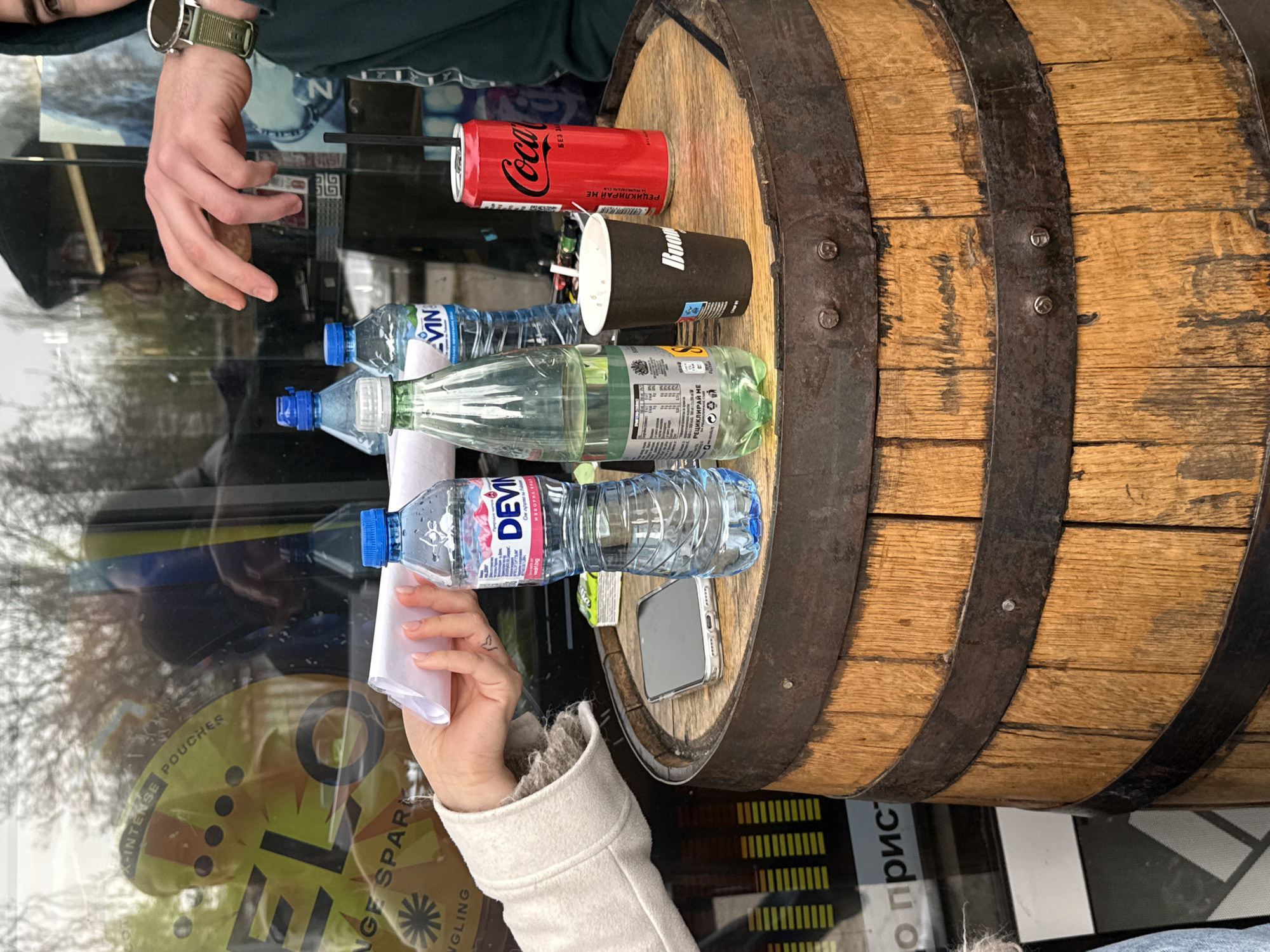 The image shows a wooden barrel, possibly used as a makeshift table, placed in what appears to be a public space in Sofia, Bulgaria. The location can be inferred from the Cyrillic writing on the Coca-Cola can and the packaging of the water bottles. On the barrel's surface are several items: three bottles of water (one "Devin" and two "Vin"), a Coca-Cola can, a disposable coffee cup with "Boo" written on it, and a smartphone. Two hands are visible, one holding a napkin near the "Devin" water bottle, and another reaching towards the objects on the barrel. The background shows a reflection of the outdoor environment on a glass surface, likely a shop window. The focus is on the barrel and the objects placed on it.