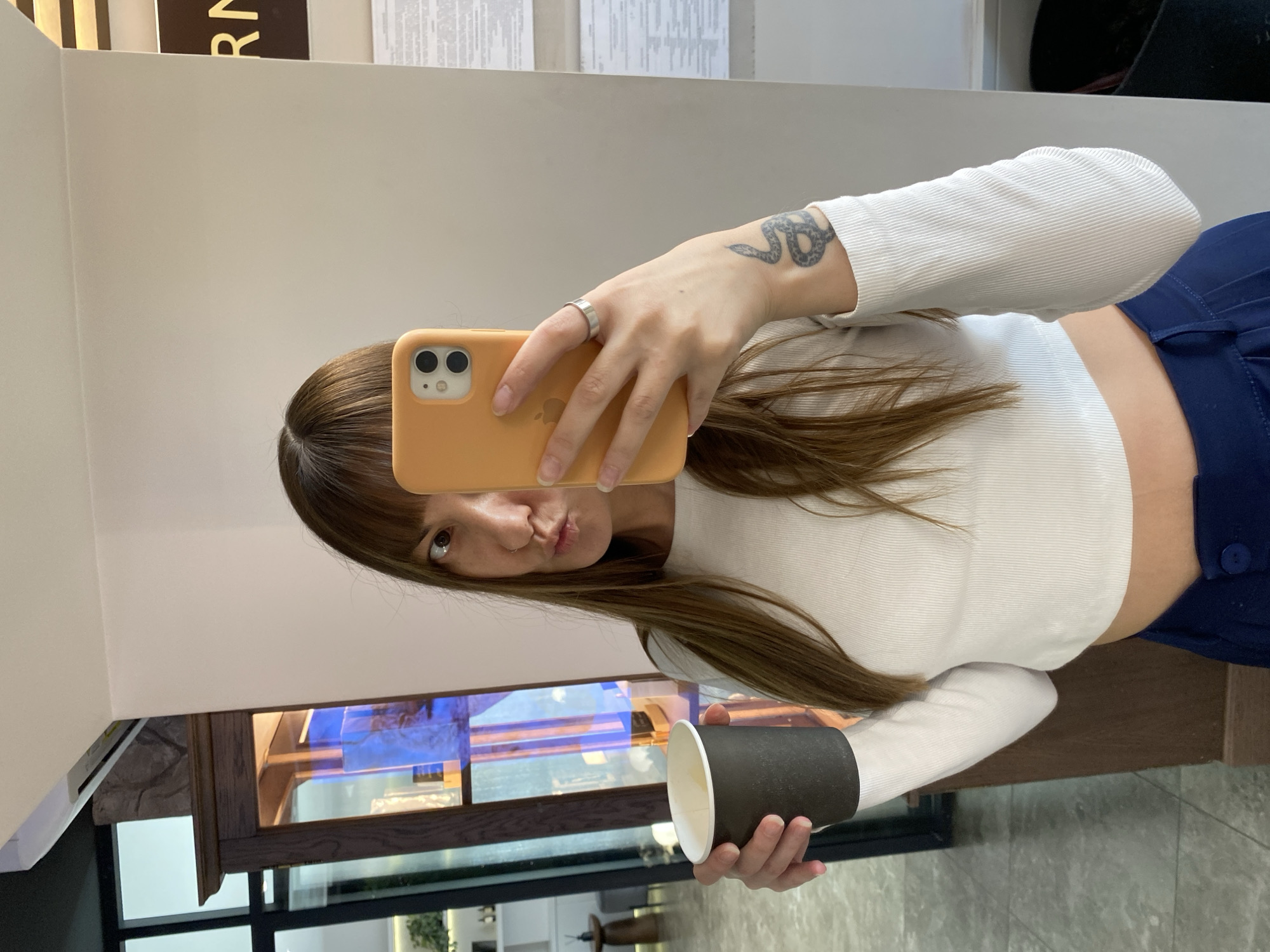 A person is taking a selfie while holding a cup. The person has long brown hair, is wearing a white long-sleeved crop top, and blue pants. A silver ring is on one of their fingers, and a snake tattoo is on their left wrist. The cup is black on the outside and white on the inside. The background shows a display case with blue light reflecting inside, and a marble-like floor. The location is unidentified and cannot be confirmed as Votkinsk, Russia from the image.