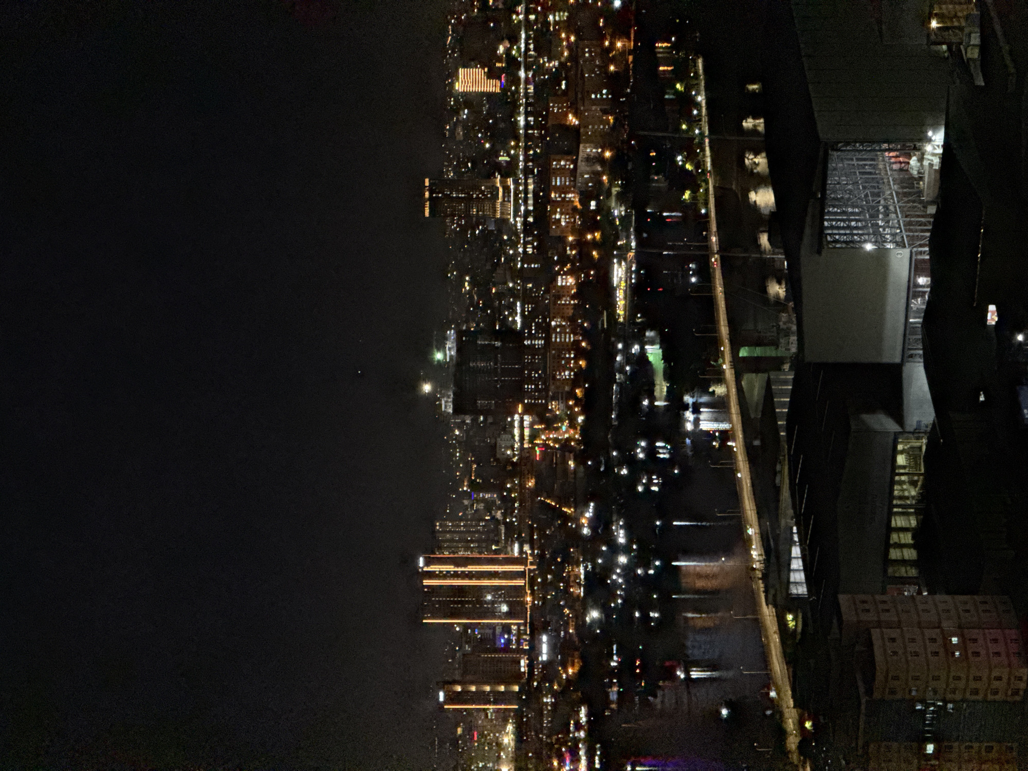 The image is a night-time aerial view of a city, likely Đà Nẵng, Vietnam, showing a river bisecting the cityscape. Numerous high-rise buildings are illuminated with bright lights, creating a vibrant urban landscape. A bridge spans the river, with cars visible on its surface. In the foreground, there are several low-rise structures, possibly industrial or commercial buildings, with some internal lighting. The sky is dark, with the city lights reflecting on the water's surface. The image's resolution is somewhat grainy due to the low light conditions.