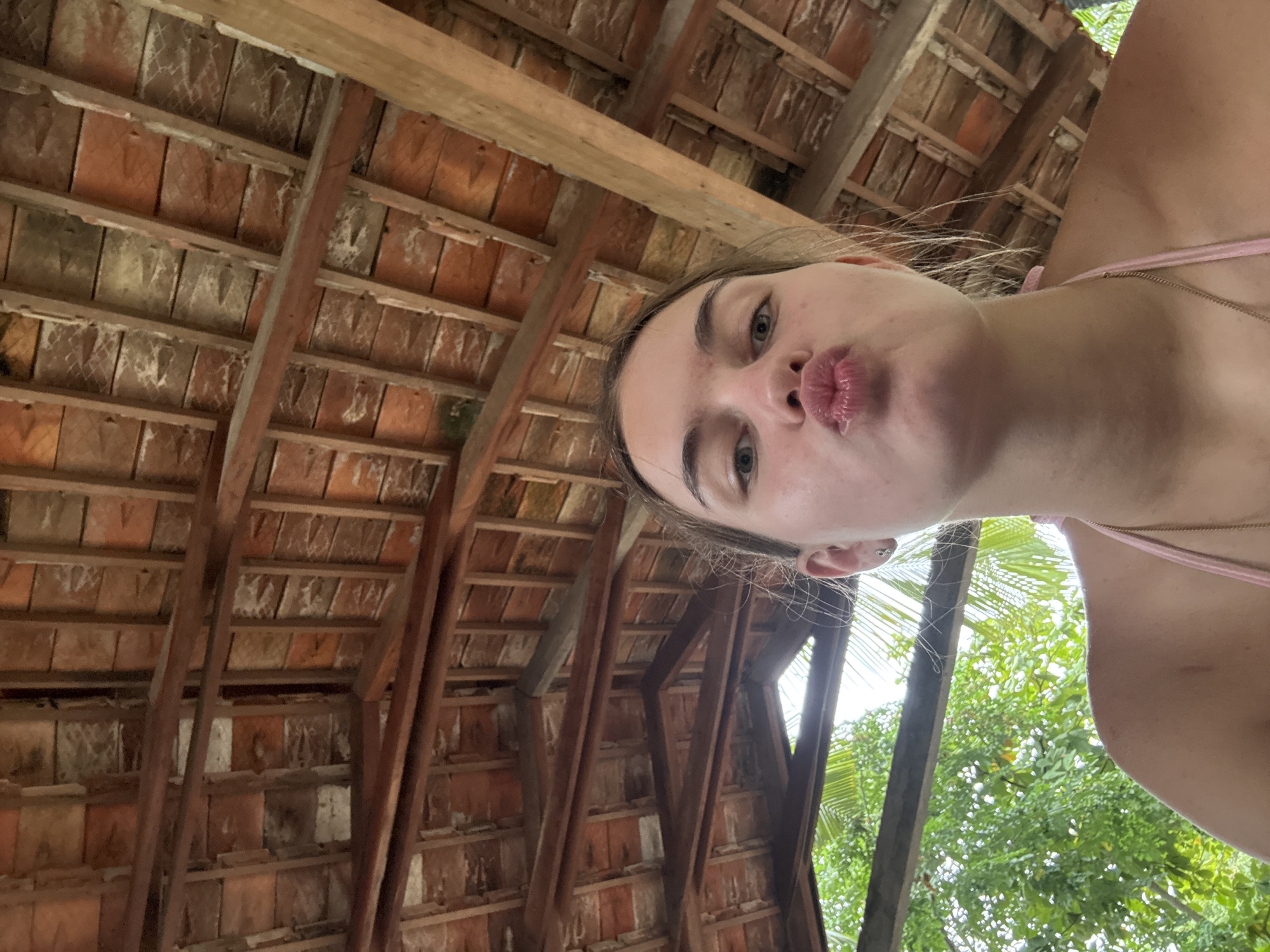 A young woman with light skin and blonde hair is pictured in a selfie, puckering her lips. She is wearing a pink bikini top. The background shows a wooden roof structure and lush green trees. The lighting suggests an outdoor, daytime setting. The image is taken from a low angle looking up at the woman. The location is not specified in the image or the provided context.