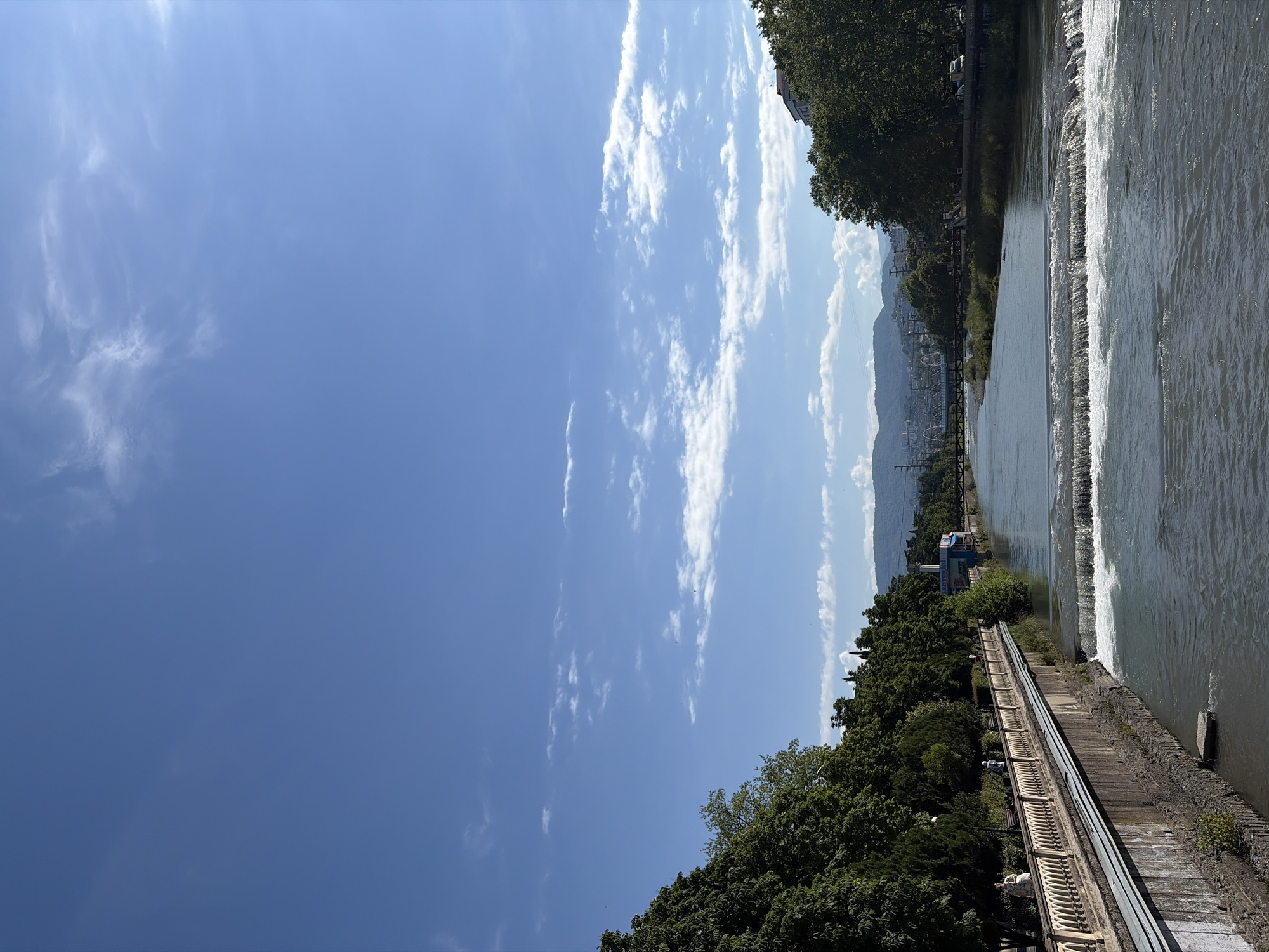 The image displays a wide-angle view of the Sochi River in Sochi, Russia, on a clear, sunny day, captured from an elevated position, likely a bridge, looking upstream.
The Sochi River dominates the lower and central portions of the frame, flowing from the distant background towards the immediate foreground. Its water is a greenish-grey color. In the middle ground, an artificial weir creates three distinct cascades, generating foamy white water across the river's width.
On the left bank, a sturdy concrete embankment supports a pedestrian promenade. This promenade features ornate, light-colored balustrades with decorative arched openings. A dense line of mature green trees, including both broadleaf and coniferous species, borders the promenade. Several individuals are present on the promenade: one person in a light-colored dress walks towards the foreground, while two other figures are visible further back, one appearing to sit on a bench and another walking. Beyond the trees in the mid-ground, a small, light-blue building with a red roof and discernible signage stands near the river.
The right bank is also thickly covered with lush green trees. Partially visible above and through the canopy of these trees are several multi-story buildings in the distant background.
The background of the scene features rolling mountains covered in vegetation, extending across the horizon. An arched bridge, appearing to be a railway or road bridge, spans the river further upstream against the mountainous backdrop. Some urban infrastructure, including power lines and poles, is discernible against the lower slopes of the mountains.
The sky, occupying the upper half of the image, is a vivid, clear blue. Scattered, wispy white clouds are distributed across the expanse, with some larger, puffier cumulus clouds visible closer to the horizon on the right. The bright sunlight indicates daytime conditions, casting distinct shadows.