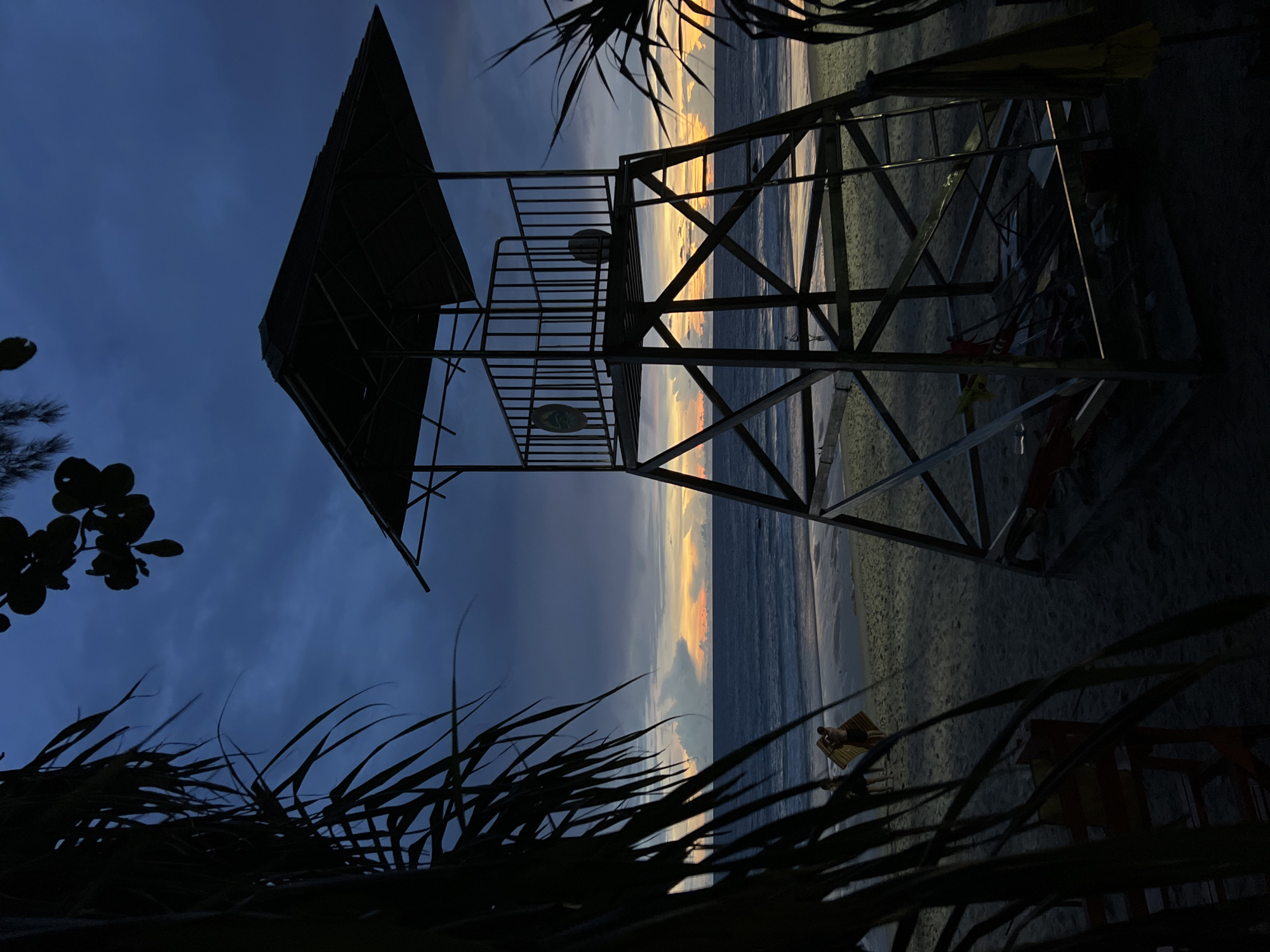 A high-angle, full shot captures a dark blue twilight sky above a beach and ocean. In the foreground, silhouetted palm fronds obscure parts of the scene on the left and right. A dark, metallic lifeguard tower stands prominently in the mid-ground, its open structure revealing a ladder and some scattered objects beneath it. The ocean stretches to the horizon, meeting a sky transitioning from dark blue to hues of orange and yellow where the sun has set. The wet sand of the beach reflects the dim light. Two figures are visible in the distance on the beach, and a red lifeguard chair is partially visible in the lower left foreground, also in silhouette.