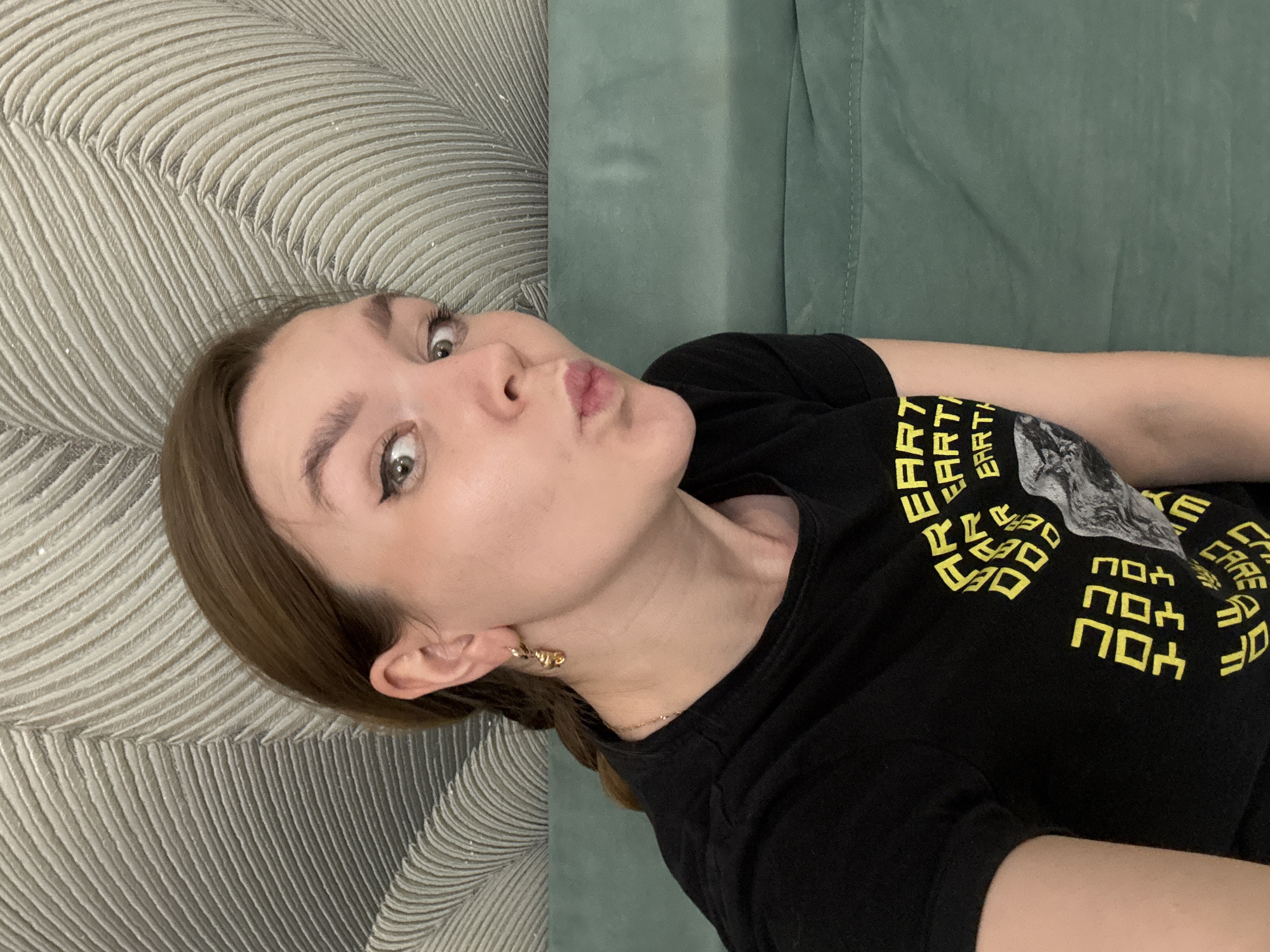A young woman with light brown hair and fair skin lies on a patterned grey chaise lounge or cushion, possibly in an indoor setting. She is wearing a black t-shirt with yellow text and a graphic print. The woman has noticeable eyeliner and is puckering her lips in a kiss-like expression. She also wears gold earrings. The background consists of the chaise lounge or cushion and a green upholstered surface. There is no visible indication of the location being Izhevsk, Russia.