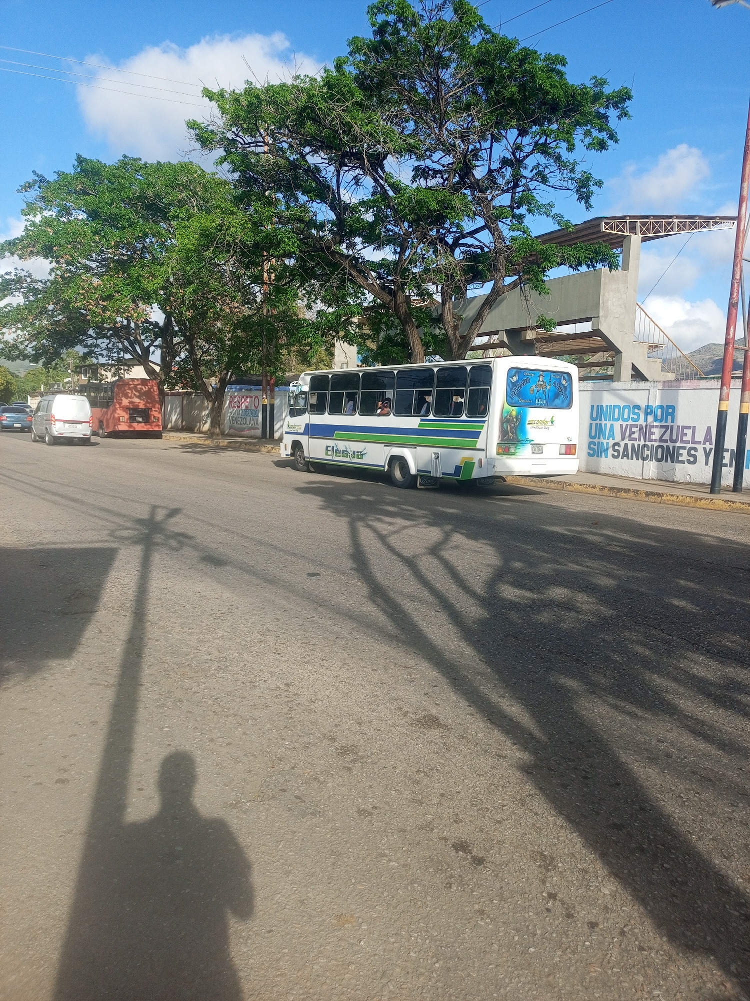 In Altagracia, Venezuela, a white bus with green and blue stripes is parked on a street. The bus has "El Conquistador" written on its side. Behind the bus, a wall features a mural with the text "UNIDOS POR UNA VENEZUELA SIN SANCIONES Y EN PAZ". To the left of the bus, a red truck and a white van are also parked. The street is paved, and shadows of trees and a person are cast across it. The sky is blue with scattered clouds.