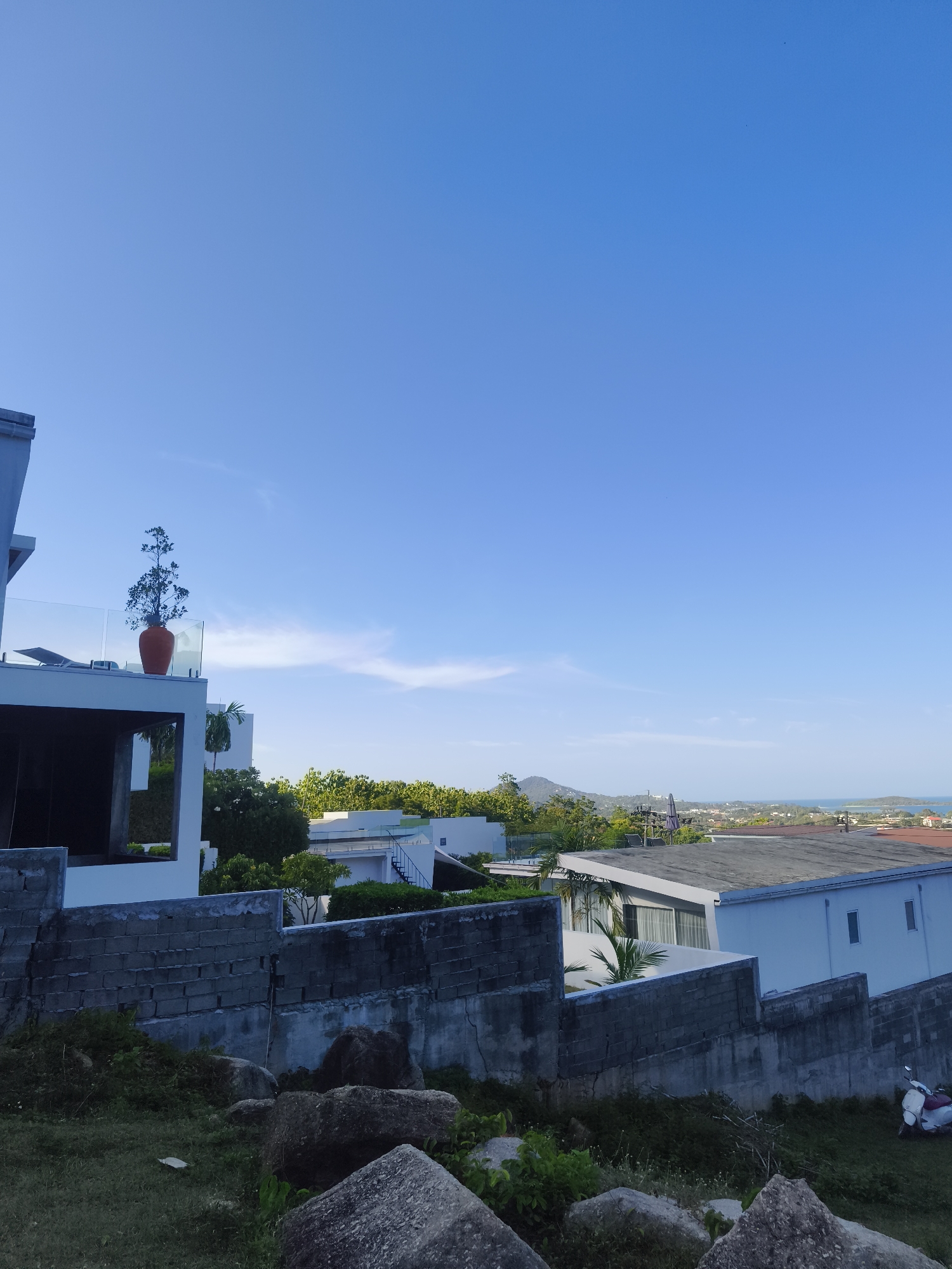 The image captures an elevated view of a modern residential area on Ko Samui, Thailand, under a clear blue sky with sparse clouds. In the foreground, large boulders and overgrown grass suggest a natural, slightly unkempt terrain. A rough, grey concrete block wall runs horizontally, serving as a boundary.
Beyond the wall, the scene transitions to a more manicured environment with several white, contemporary buildings. One prominent building on the left features a balcony with a glass railing and a tall, slender tree in an orange pot. Another building to the right has a flat, dark roof and large windows. Further in the distance, more white structures are visible, interspersed with lush green trees and vegetation. The landscape slopes downwards towards a hazy horizon where the ocean meets the land, with islands or landmasses visible in the distance. A white scooter with a red seat is parked on the grassy area near the concrete wall on the right side of the image. The overall impression is one of a modern villa complex situated on a hillside overlooking the sea in a tropical locale.