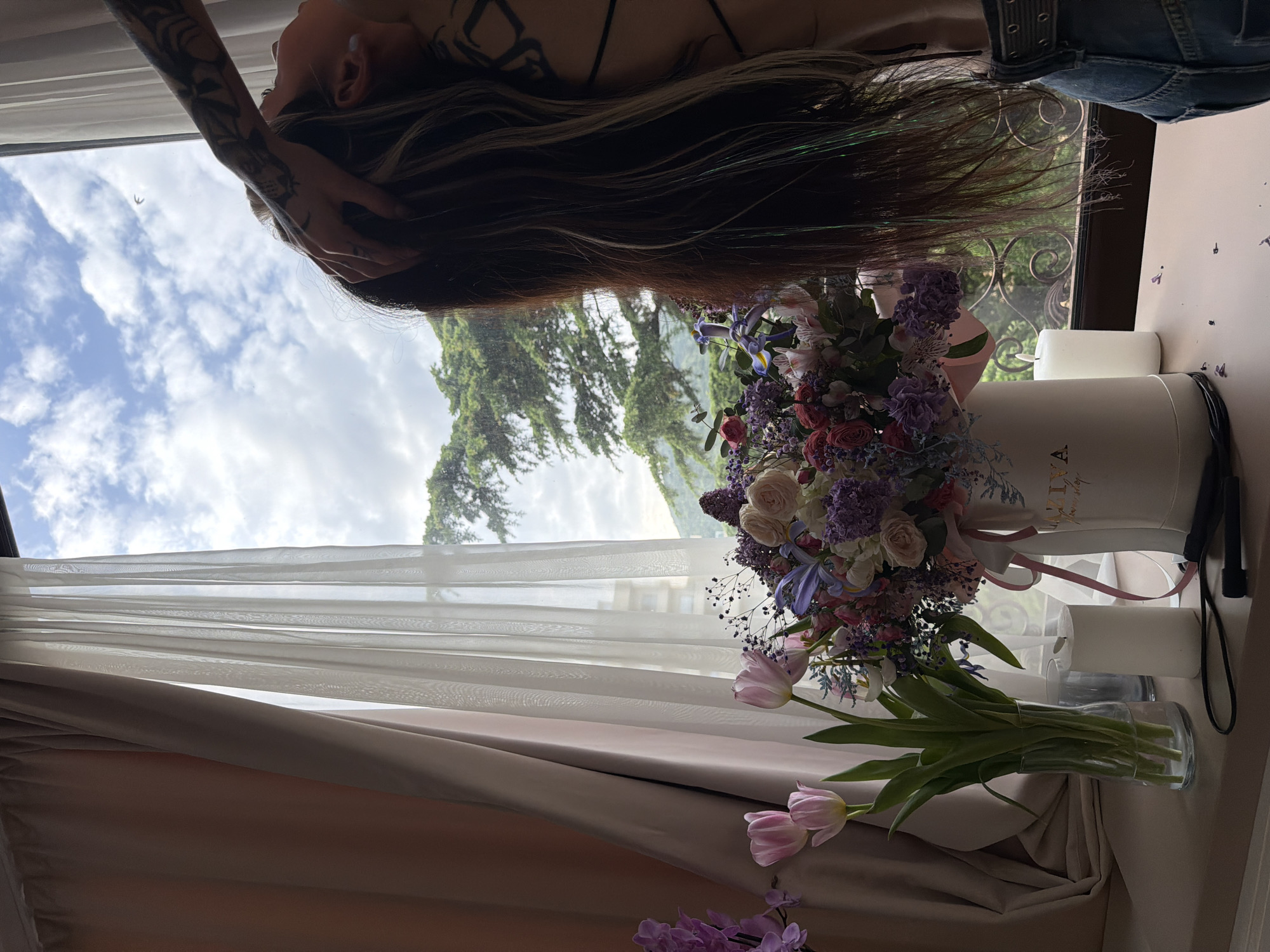 The image captures an indoor scene by a large window, offering a view of the outdoors. In the foreground, a person is lying down, with their head near the window. Their long, dark hair, with subtle greenish and lighter streaks, cascades downwards. Tattoos are visible on their arm and potentially shoulder/back. They appear to be wearing a dark top or bra and blue jeans.
On the right side of the image, two floral arrangements are present. A large, ornate bouquet is housed in a tall, cylindrical white box. This bouquet features a variety of flowers, including pink and white roses, purple and blue irises, clusters of smaller purple flowers (possibly hydrangeas or similar), and delicate baby's breath. Text on the white box clearly reads "FLOWER SHOP" and possibly a brand name like "LIZIYA." Below this, a clear glass vase holds a bunch of fresh pink tulips, and at the very bottom, some purple flowers (possibly orchids) are visible. Two white cylindrical objects, resembling candles, are placed behind the large flower box.
The window reveals a cloudy daytime sky with patches of blue, and a lush green landscape featuring tall conifer trees. A distant, less distinct building can also be seen. Sheer white curtains and heavier beige/taupe curtains hang by the window, framing the view. The overall impression is one of serene contemplation or relaxation in a room adorned with fresh flowers, overlooking a natural environment. The location is Budva, Montenegro.
