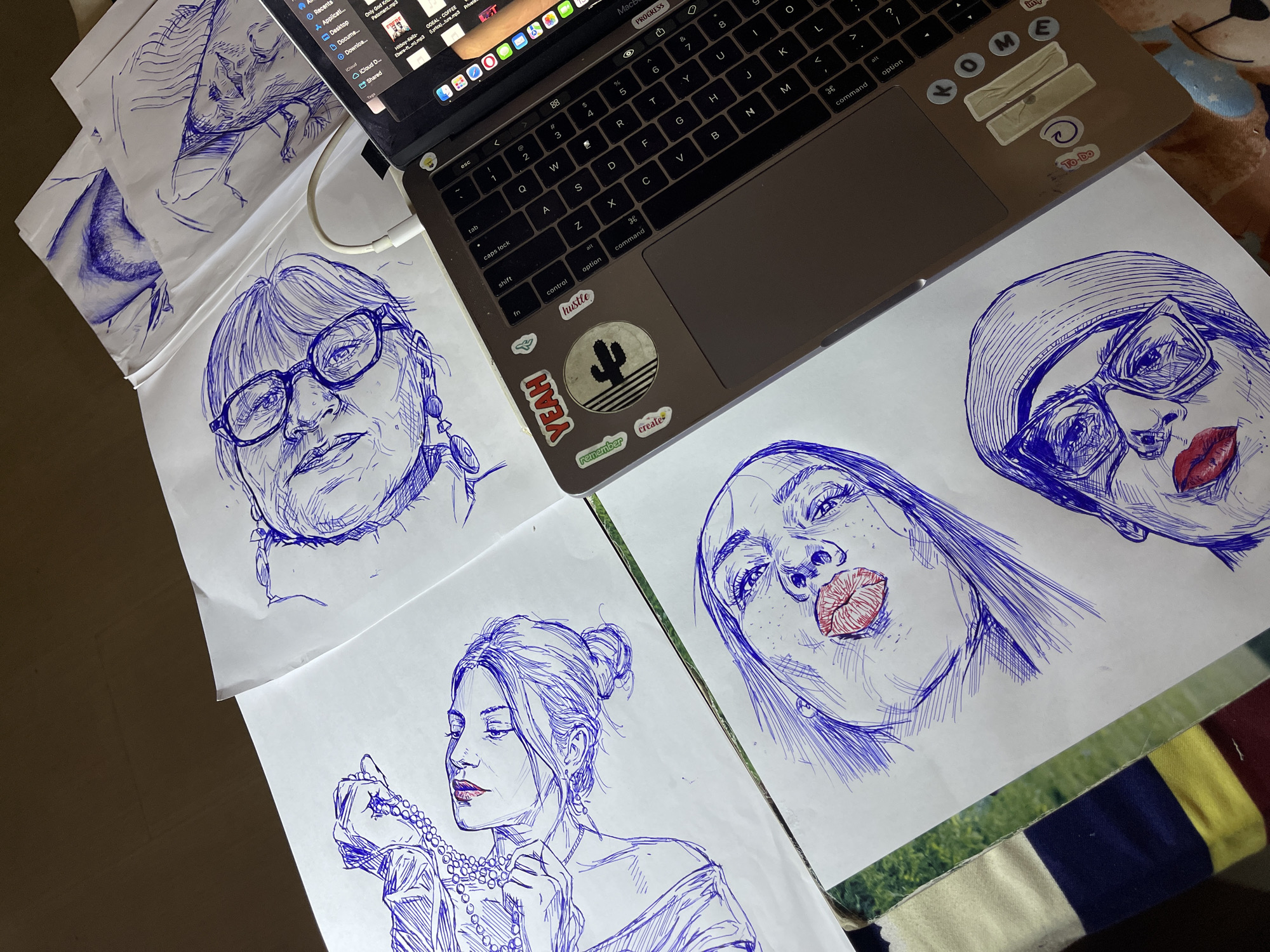 A top-down view of a workspace shows several blue ink pen drawings of women's faces on white paper scattered around a laptop. The drawings vary in completion and pose. One depicts a woman with glasses and braided hair. Another shows a woman with eyes closed, holding a string of pearls. A third drawing focuses on a woman's face with freckles and red lipstick, looking upwards. A fourth drawing shows a woman wearing glasses and a headscarf, also with red lipstick. The laptop, positioned on the right side of the frame, is open and its screen displays a colorful user interface, likely a software application. Several stickers are affixed to the laptop's casing, including a "YEAH" sticker and a sticker with a cactus graphic. The scene is illuminated by an overhead light source, casting soft shadows. The background is a dark, indeterminate surface, suggestive of a table or floor. The presence of the laptop and the scattered drawings imply an artistic or creative process. The image is devoid of any discernible landmarks or clues that would associate it with the city of Jos, Nigeria.