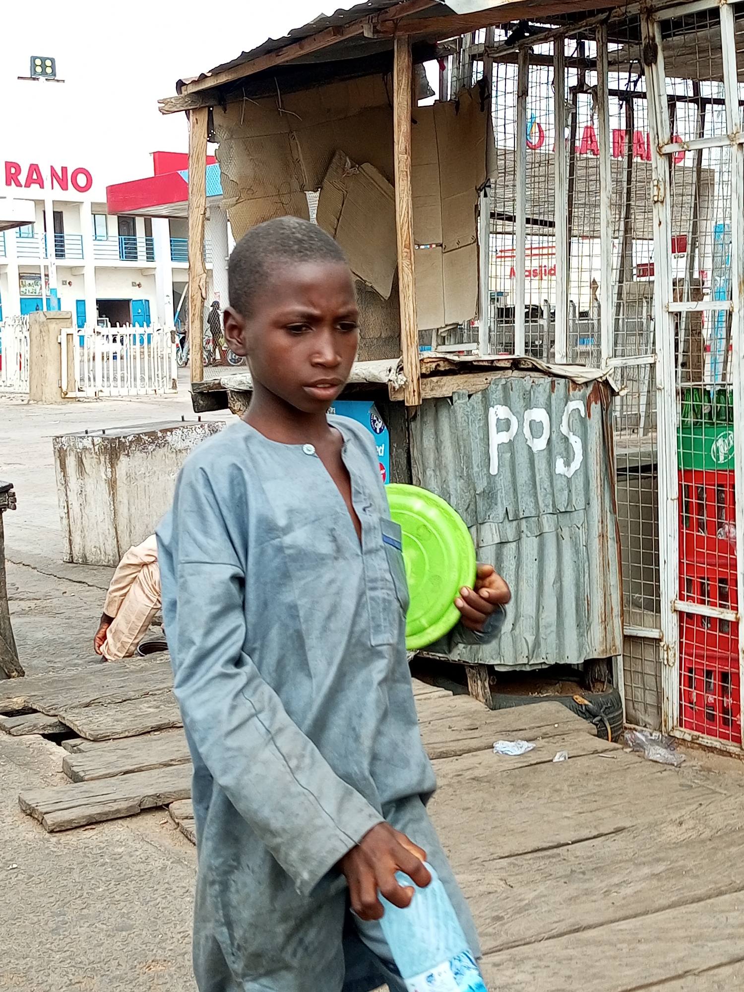 The image depicts a street scene in an urban environment, likely in a market or commercial area. In the foreground, a young Black male stands facing slightly to the right, wearing a light blue, long-sleeved garment that appears to be a tunic. He is holding a green circular object in his left hand and a clear plastic bottle of water in his right hand. His gaze is directed downwards and to the right.
Behind the boy, a corrugated metal structure with the letters "POS" painted in white is visible. To the right of this structure, a metal cage encloses shelves stacked with red crates, some of which contain green bottles. Further in the background, a building with red and blue accents and the text "RANO" in red letters is present. A white fence is in front of this building. The ground is covered with wooden planks and concrete. In the mid-ground, another individual, possibly male, is seen crouching or kneeling. The scene appears to be during daylight.