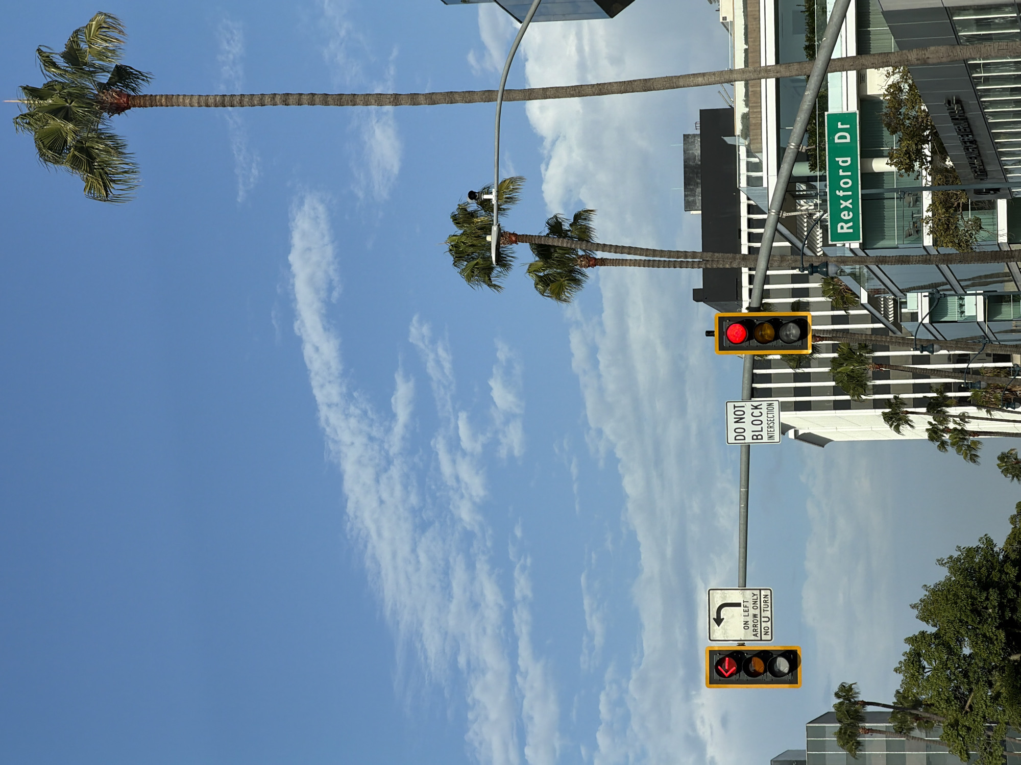 A tall palm tree dominates the left side of the frame, its fronds reaching towards a clear blue sky with scattered wispy clouds. In the background, modern buildings with reflective glass windows rise up. A street sign with green lettering reads "Rexford Dr."
In the middle ground, a traffic light displays a red signal. Below it, a sign indicates "DO NOT BLOCK INTERSECTION." Further down, another traffic light shows a red signal, and a regulatory sign features a U-turn arrow with the text "ON LEFT ARROW ONLY NO U TURN."
The scene appears to be a bright, sunny day in a city environment, likely Beverly Hills, given the context provided. The presence of palm trees and upscale architecture suggests a pleasant, possibly warm, climate. There are no people visible in the image, and the focus is on the urban landscape and traffic control elements. The image is oriented vertically, with the sky at the top and the street level at the bottom.