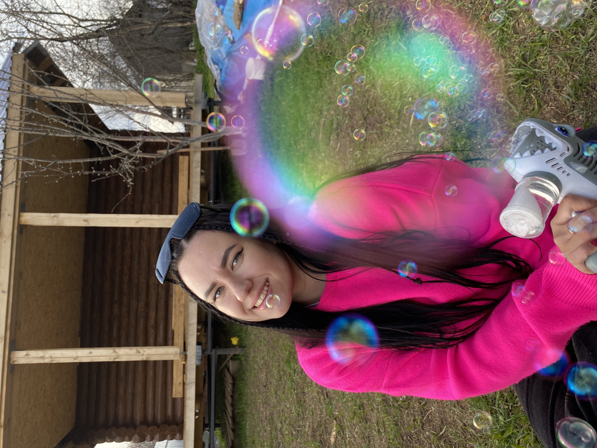 A young woman with long dark hair, wearing a vibrant pink long-sleeved top and dark clothing, smiles cheerfully at the camera while engaging in a playful outdoor activity. She holds a grey and white shark-shaped bubble gun in her right hand, from which numerous iridescent soap bubbles are being emitted. These bubbles, reflecting rainbow colors, float around her. Sunglasses are perched on her head.
The scene is set outdoors in what appears to be a backyard or rural area in Chaikovsky, Russia. The woman is positioned on green grass, suggesting springtime or summer. In the background, there is a wooden structure, possibly a shed or an outbuilding under construction, featuring light wooden beams and darker timber siding. Bare tree branches are visible against a bright, possibly overcast, sky. The image captures a moment of joy and lightheartedness during the daytime. No visible text is present in the media file.