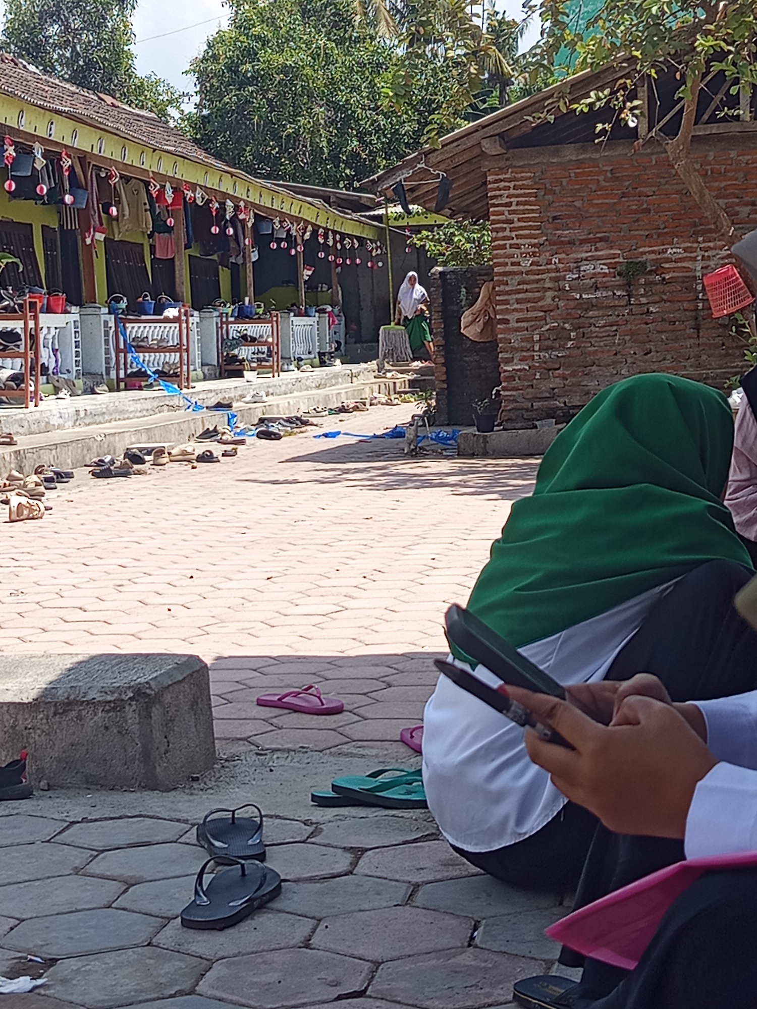 The image depicts an outdoor scene in Lumutan, Indonesia, likely at a religious or educational institution. In the foreground, several pairs of footwear, including flip-flops and sandals, are scattered on a brick-patterned ground. A person wearing a green hijab and white clothing is seated on the ground, holding a stylus over a tablet. Another individual's hands are visible in the lower right, also holding a stylus near a device, with a pink fabric draped over their lap.
The background reveals a building with a green facade and a tiled roof, adorned with decorative elements and hanging items that appear to be bags and garments. Shoe racks are positioned along the building's front. Further back, a brick structure with a wooden roof is visible, with foliage and trees surrounding the area. Two women, one in a white hijab and another in a green garment, are walking near a doorway to the left of the brick building. The overall environment suggests a sunny day with a bright sky.