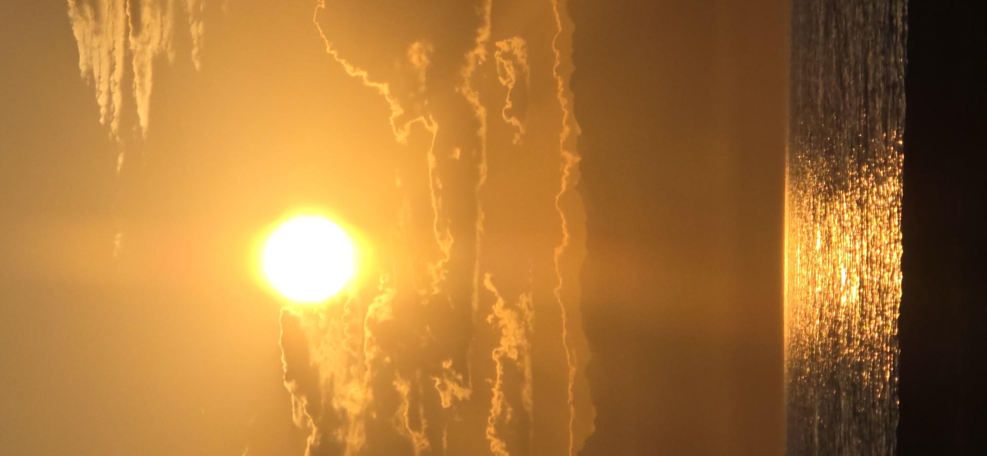 The image depicts a vibrant sunset over a body of water, likely the Issyk-Kul lake in Boz Beshik, Kyrgyzstan. The sun, a brilliant white orb, is partially obscured by scattered, golden clouds, casting a warm, orange glow across the sky. Rays of sunlight pierce through the atmosphere, creating a soft, diffused light. Below the sky, the water reflects the fiery hues of the sunset, creating a shimmering, golden pathway across its surface. The foreground is dominated by a dark, silhouetted shore, providing a stark contrast to the luminous sky and water. There are no people or visible objects other than natural elements. The environment is serene and tranquil, captured during the twilight hours.