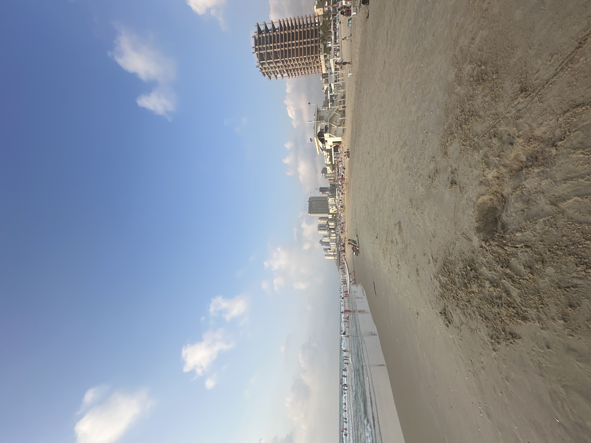 The image depicts a wide sandy beach in Bat Yam, Israel, under a clear blue sky with scattered white clouds. The foreground is dominated by sand, with some ripples and footprints visible. In the middle ground, the gentle waves of the sea wash onto the shore, where numerous people are seen swimming, walking, and relaxing. Several red flags mark boundaries on the beach and in the water. Further back, a lifeguard station with a white structure stands prominently. Beyond the beach, the skyline of Bat Yam is visible, featuring a collection of modern buildings and a tall, unfinished skyscraper. A few beach chairs and umbrellas are scattered on the sand, and some individuals are seated in chairs. The overall scene conveys a relaxed atmosphere of a popular beach destination.