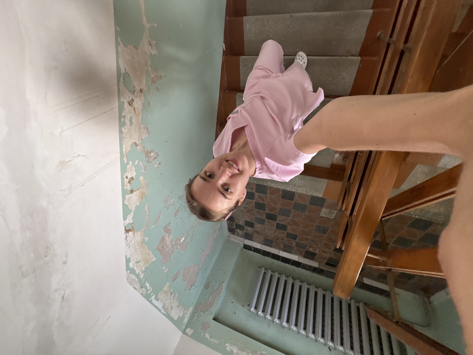 A woman in a pink medical uniform stands on a stairwell, holding a camera to take a selfie. The stairwell is in a state of disrepair, with peeling green paint on the walls and ceiling, and worn tiled flooring. A radiator is visible on the left wall. The woman is looking directly at the camera with a smile. The scene appears to be in an older building, possibly in Chaikovsky, Russia, given the context.