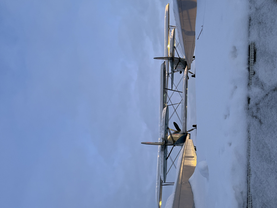 Two biplanes covered in snow stand adjacent to a snow-covered rooftop. The planes, arranged vertically in the frame, feature propellers and wings coated with snow. The pale blue sky forms the backdrop, providing a cool contrast to the white snow. The rooftop, lined with a dark, textured edge, displays scattered footprints in the snow.