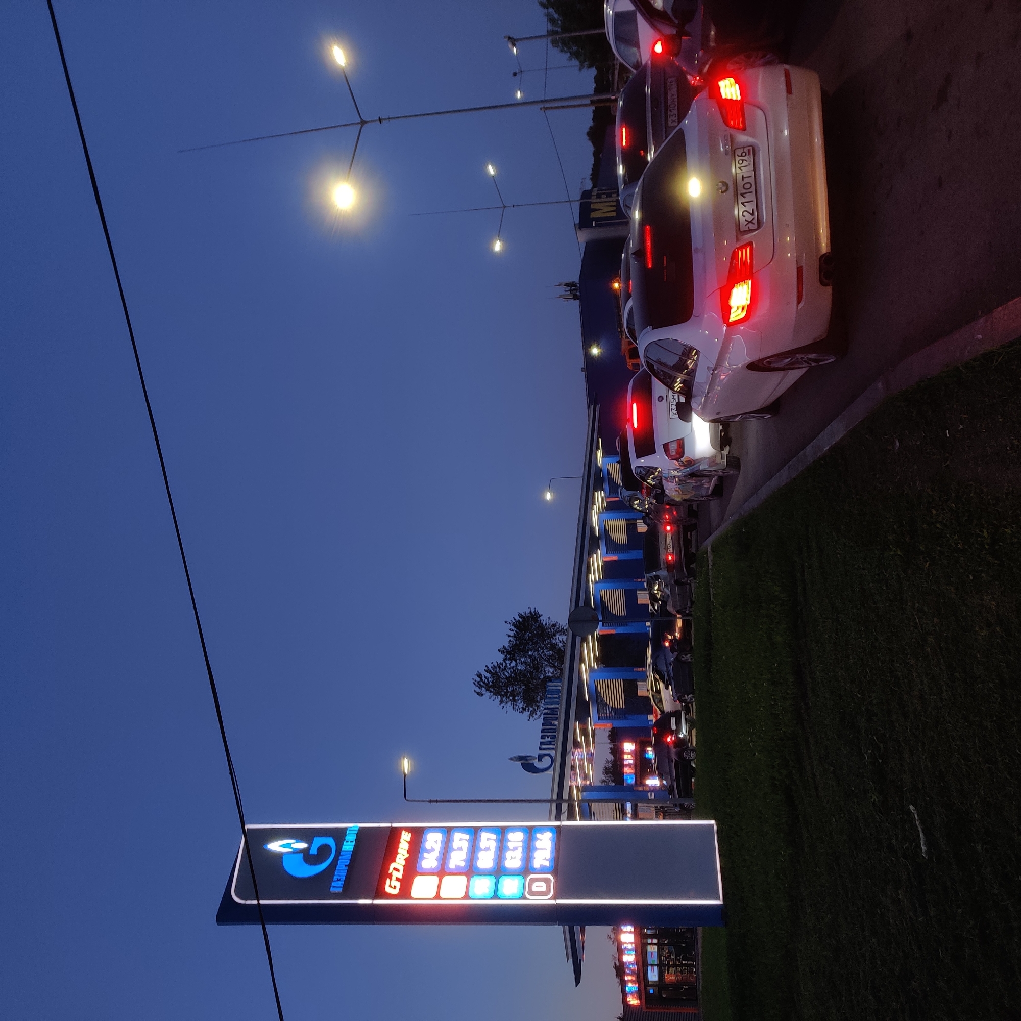 A long line of cars is waiting at a gas station at dusk. The gas station has a tall, illuminated sign with the Gazprom logo and fuel prices displayed. The sky is dark blue, indicating either early morning or late evening. The cars are stopped at a pump, and some have their headlights on. The scene suggests a busy time at the gas station, possibly due to high fuel prices or a rush hour. The overall atmosphere is calm, despite the queue.