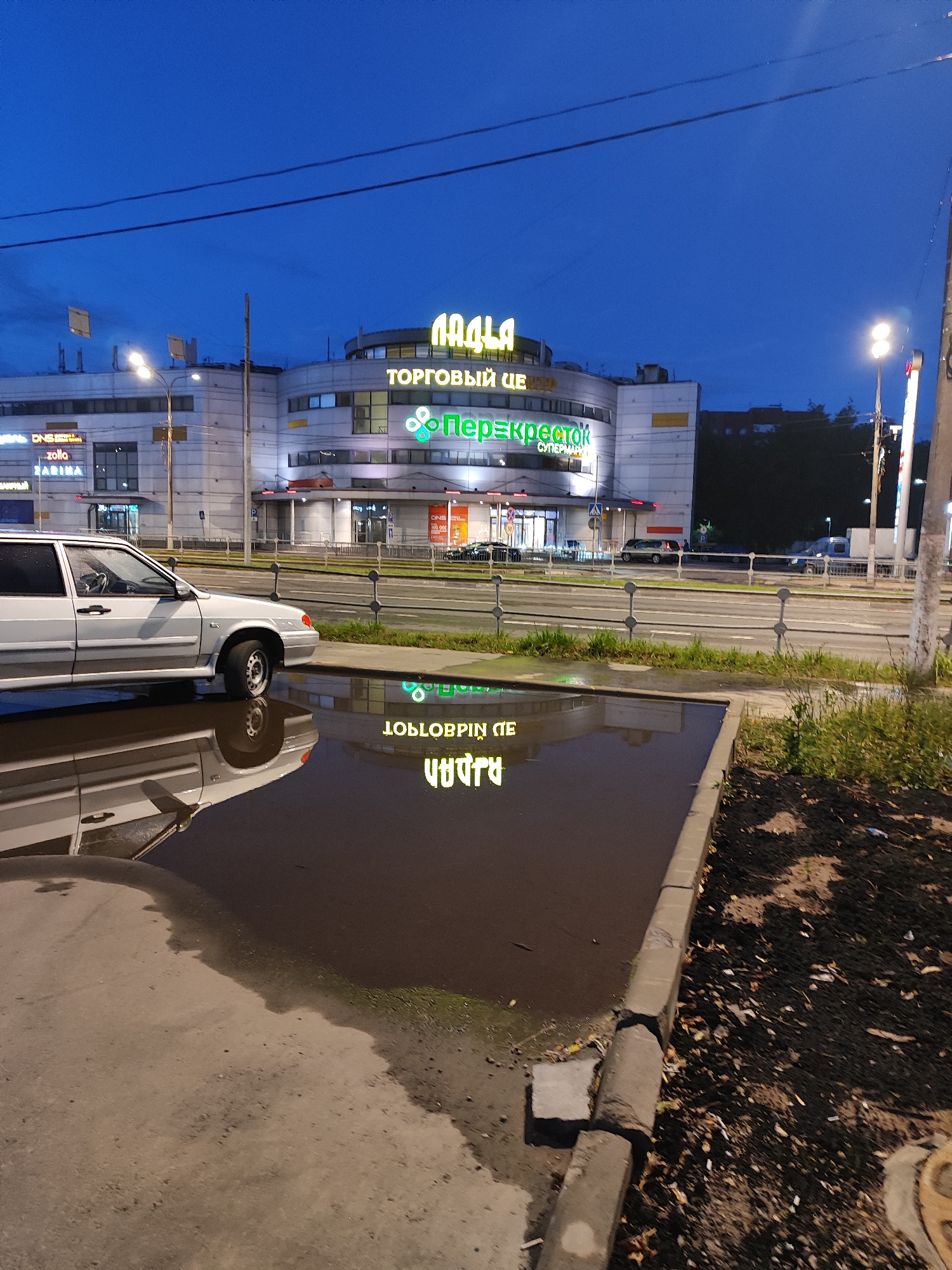 A long-shot, eye-level, outdoor photograph taken at dusk shows a silver sedan parked next to a large puddle of water on an asphalt surface. The sedan is positioned on the left side of the frame, facing right. The puddle occupies the central and lower portions of the image, reflecting the lights of a multi-story building and the sky. The building, located in the background, features illuminated signage in green and yellow, with the words "ТОРГОВЫЙ ЦЕНТР" (Shopping Center) and "Перекрёсток" (Perekrestok) clearly visible. The sky above is a deep blue, characteristic of twilight. The scene is urban, with a road and sidewalk visible in the middle ground. The ground around the puddle is covered with dark soil and some scattered leaves. The overall atmosphere is subdued due to the low light conditions.