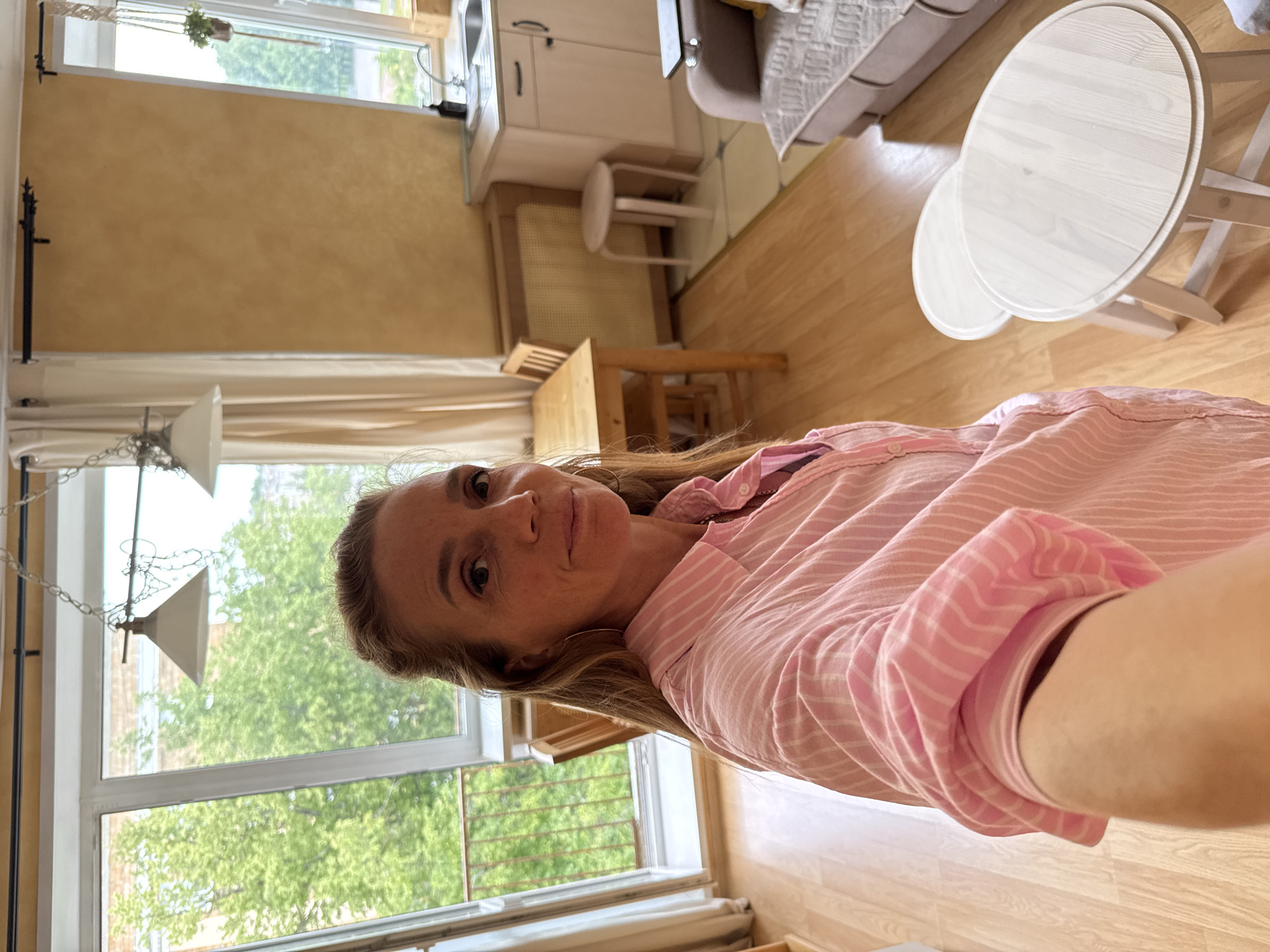 The image is a selfie taken by a woman in what appears to be a small apartment or studio. She is wearing a pink and white striped button-down shirt and large hoop earrings. Her hair is pulled back. The background shows a living space with a window overlooking greenery, a small dining table with chairs, a kitchen counter with cabinets and a sink, and a sofa. There are also two round, light-colored stools in the foreground. The lighting suggests it is daytime. The setting is likely an urban residential area. No specific landmarks or indicators place this scene definitively in Veliky Novgorod, Russia, beyond the prompt's assertion.