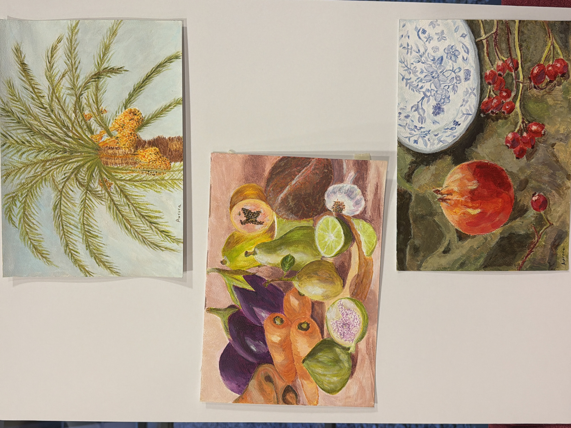 The image displays three framed paintings hung on a white wall. The top painting is of a palm tree with clusters of ripe dates. The middle painting is a still life featuring eggplants, carrots, figs, pears, limes, papaya, coconut, and garlic. The bottom painting shows a pomegranate, a decorative plate with a blue floral pattern, and a branch with red berries. All paintings appear to be signed "Aurora". The paintings are displayed in a gallery or exhibition space in Elche, Spain.