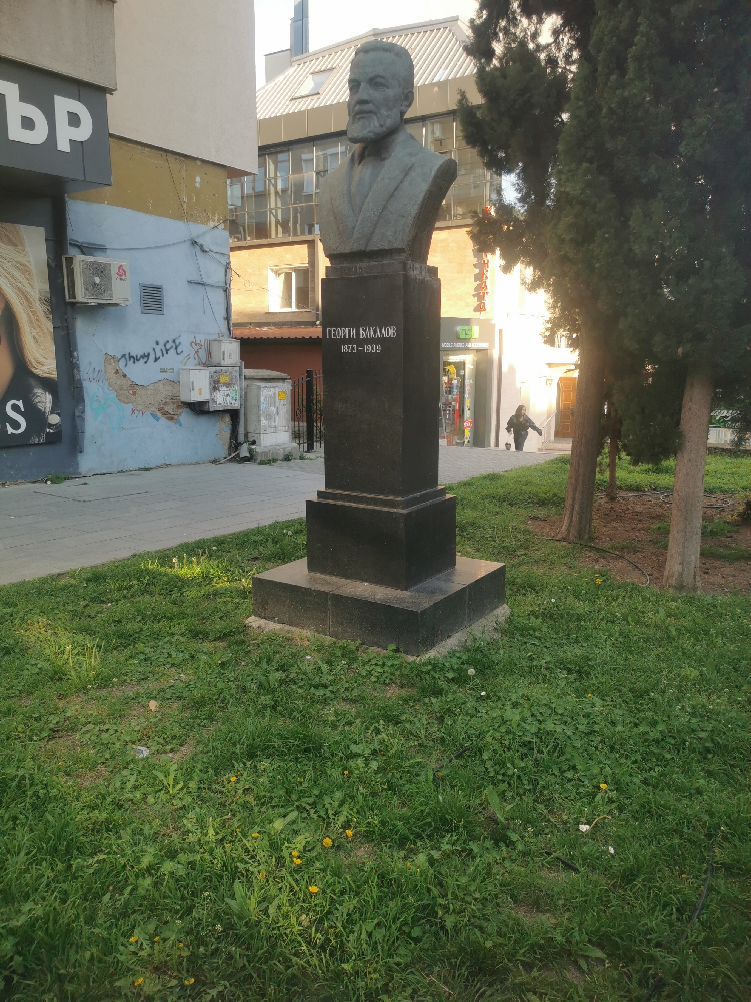 A bronze bust of Georgi Bakalov (1873-1939) stands on a tall, dark granite pedestal in a grassy urban park. The bust depicts a man with a full beard and mustache, wearing a suit jacket and collared shirt. The pedestal is inscribed with his name in Cyrillic, "ГЕОРГИ БАКАЛОВ", and his birth and death years, "1873-1939".
The bust is positioned on a patch of somewhat overgrown grass, scattered with dandelions, adjacent to a paved sidewalk. Several tall, coniferous trees are planted around the bust and along the sidewalk to the right. In the background, various buildings frame the scene under bright, sunny conditions, suggesting late morning or afternoon.
To the left, a building features a large sign with partial Cyrillic text, "ър". Below this, a light blue wall shows cracked paint and "Thug LIFE" graffiti, along with two visible air conditioning units. Further back, more contemporary buildings with glass windows and pitched roofs are visible. To the right, along the sidewalk, a shop with a green sign is visible, advertising "MOBILE PHONES AND ACCESSORIES." A lone person is seen walking past this shop in the distance. The overall setting is an active city environment, incorporating green spaces, historical monuments, and commercial areas.