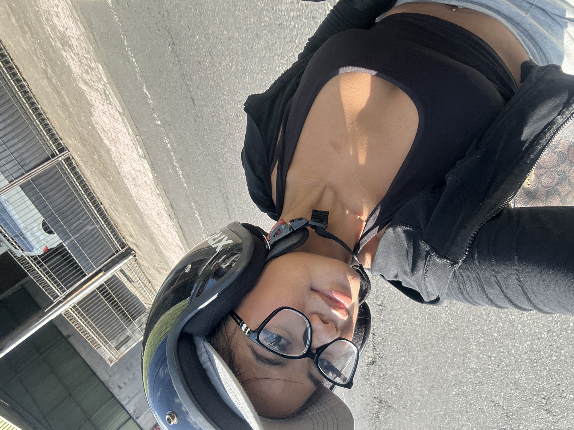 A young woman wearing a black helmet, glasses, and a black tank top is taking a selfie while on a scooter. She is wearing a grey baseball cap under her helmet. She is in an outdoor environment with a paved road and a fence in the background. The image appears to be taken in the city of Sukawati, Indonesia, as indicated by the presence of a scooter, which is a common mode of transportation in the region, and the tropical climate suggested by the lighting and attire.