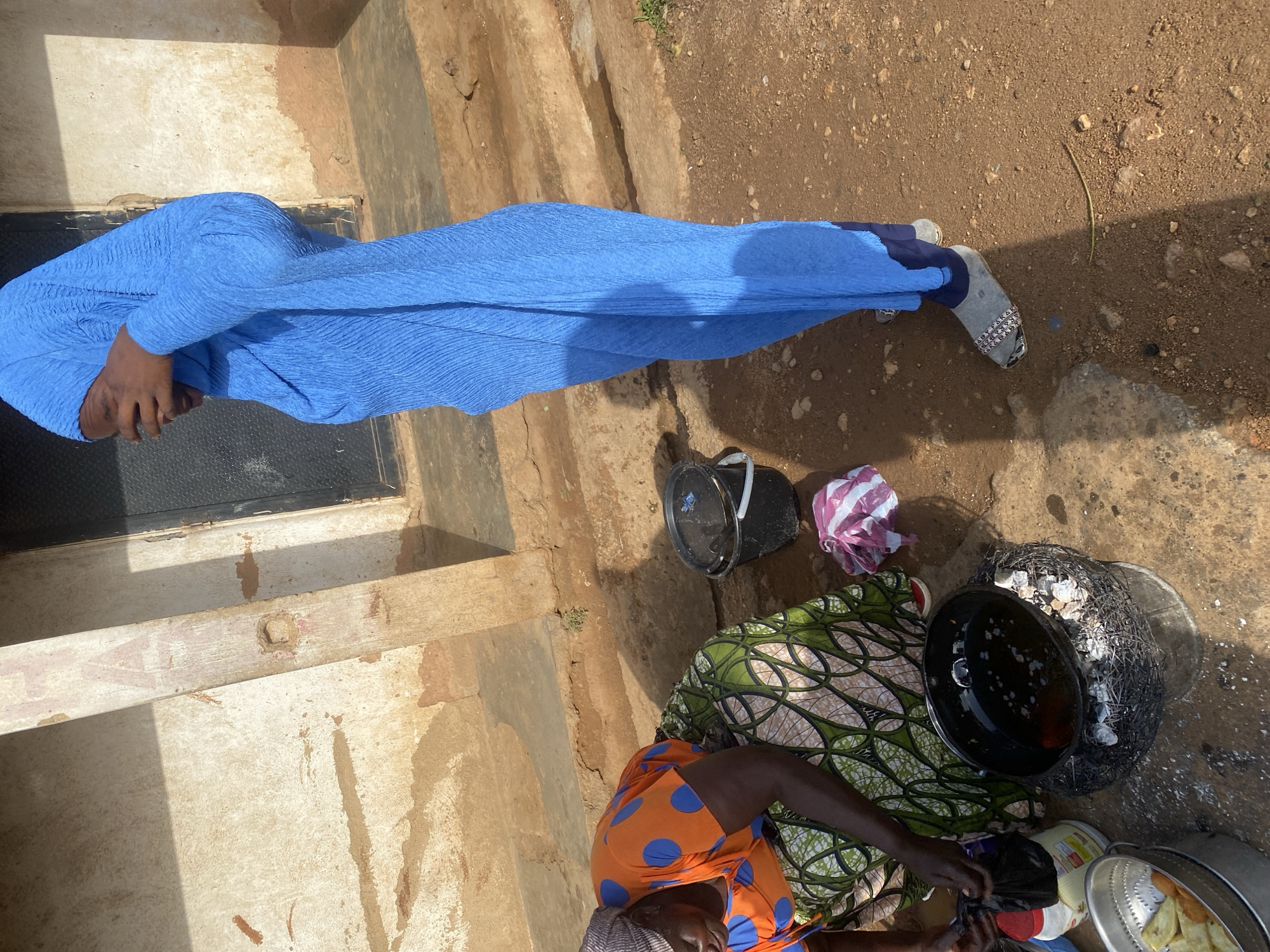 The image shows a street scene in Jos, Nigeria. In the foreground, a person is preparing food over an open fire. A large black pot is suspended over a makeshift stove made of wire mesh and burning coals. The pot contains hot oil, and pieces of food are being fried. Next to the stove are a metal colander filled with fried food, a plastic bottle, and a black plastic bag.
To the left, another person is seated, wearing an orange shirt with blue polka dots and a patterned green and white skirt. They are wearing a headscarf and appear to be assisting with the food preparation.
In the background, a person wearing a blue hijab and blue trousers stands next to a building. The building has rough, textured walls and a dark metal door. The ground is covered with reddish-brown dirt, with some stones and debris scattered around. Shadows suggest that the sun is high in the sky, indicating it is likely daytime. The overall impression is a scene of everyday life and commerce on a street in Jos.