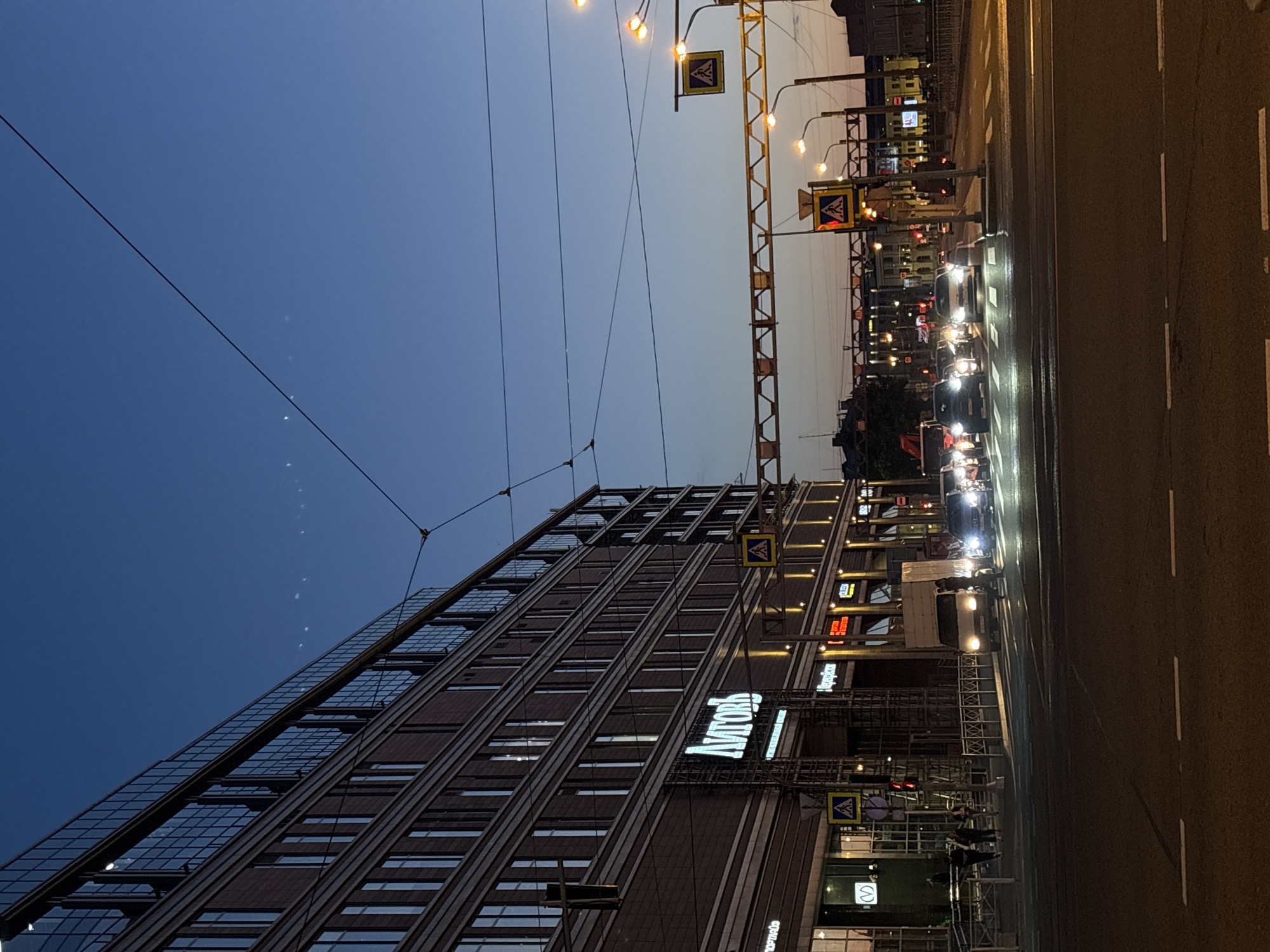 The image captures a street scene in Saint Petersburg, Russia, at dusk. A large, modern building with numerous windows lines the left side of the frame. Below the windows, a large illuminated sign reads "ЛИТОГРАФИЯ" (LITOGRAFIYA), indicating a lithography printing house or related establishment.
The street in front of the building is a busy intersection. Several cars are visible, their headlights on, as they navigate the road and crosswalks. A white truck is also present, positioned at an angle. Pedestrians are seen walking on the sidewalk and crossing the street, some illuminated by the streetlights. Traffic lights and pedestrian crossing signs are mounted on poles. Overhead, power lines and what appear to be tram wires crisscross the sky. The sky itself is a deep blue, transitioning from twilight colors. The architecture of the buildings in the background suggests a European city, consistent with Saint Petersburg. The overall atmosphere is one of urban activity as the day ends.