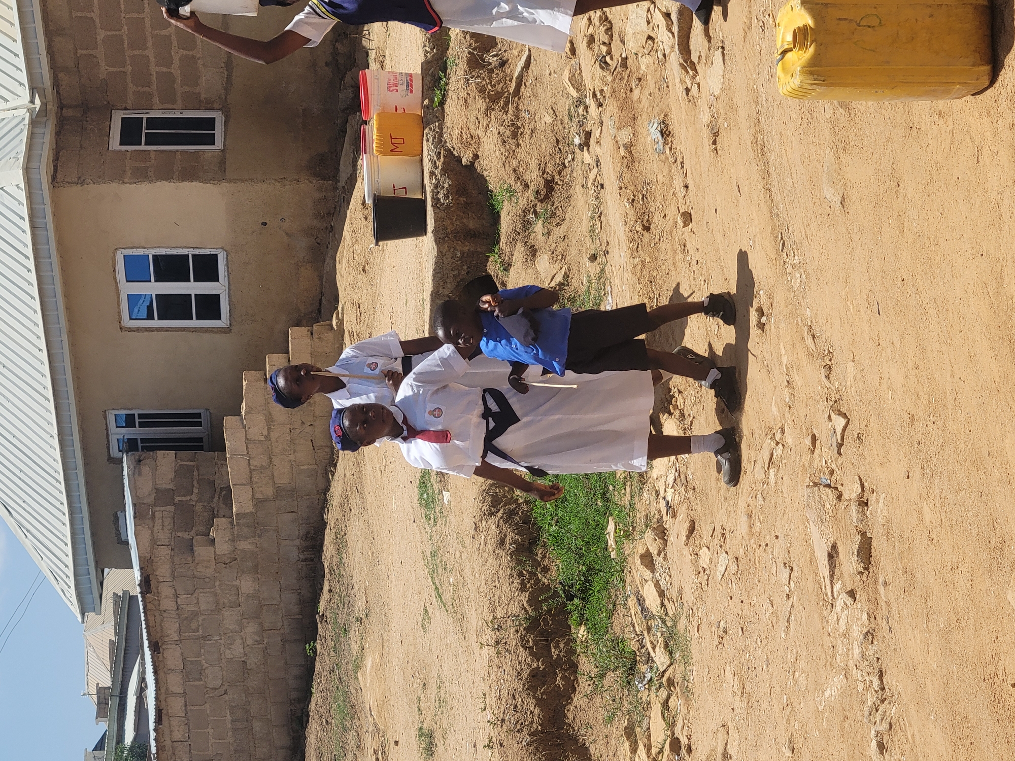 The image shows a dirt road in what appears to be a residential area of Jos, Nigeria. Several individuals are visible, some in school uniforms with a white top and a red necktie, another in a blue t-shirt and dark shorts, and another individual with a white t-shirt with navy blue accents. They are standing on the dirt road. The background includes buildings constructed with cement blocks and corrugated metal roofing, indicating a low-income urban environment. A yellow plastic container is on the far right of the image, along with three plastic buckets. The weather is clear and sunny, casting distinct shadows. The environment is dusty. The buildings appear to be in various states of completion.