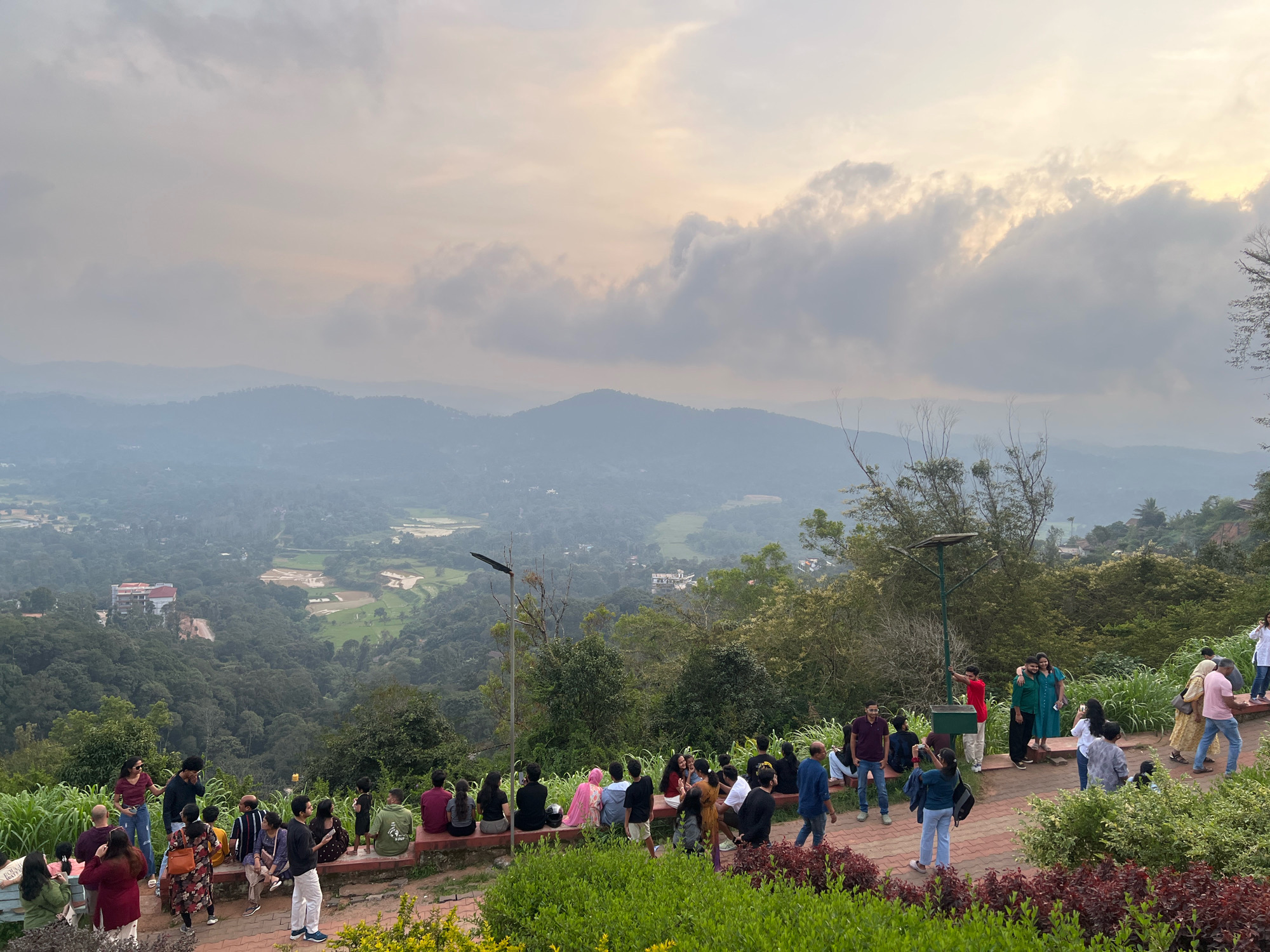 The image captures a scenic overlook with a group of people enjoying the panoramic view of the landscape surrounding Madikeri, India. In the foreground, several individuals are seated and standing on a raised viewing platform made of brick and stone, overlooking a valley. A few people are walking along a paved path. The landscape below is characterized by lush green forests, rolling hills, and some agricultural fields. In the mid-ground, there are buildings and a golf course visible. The sky is overcast with clouds, creating a soft, diffused light. The atmosphere appears relaxed and leisurely, with people engaged in conversations, taking photos, and simply admiring the vista. The overall scene suggests a popular tourist destination, likely a viewpoint offering expansive views of the Coorg region.