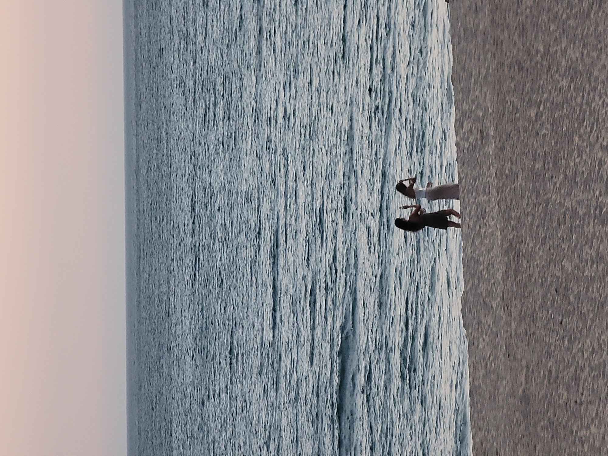 Two young women are standing on a sandy beach in Quarteira, Portugal, facing the ocean. The woman on the left is wearing a dark dress and is holding a phone to take a photo. The woman on the right is wearing a white top and a light-colored skirt, and she appears to be adjusting her hair. The sea is calm with gentle waves, and the sky is a pale pinkish-orange, suggesting it is either sunrise or sunset. The sand is a light beige color.