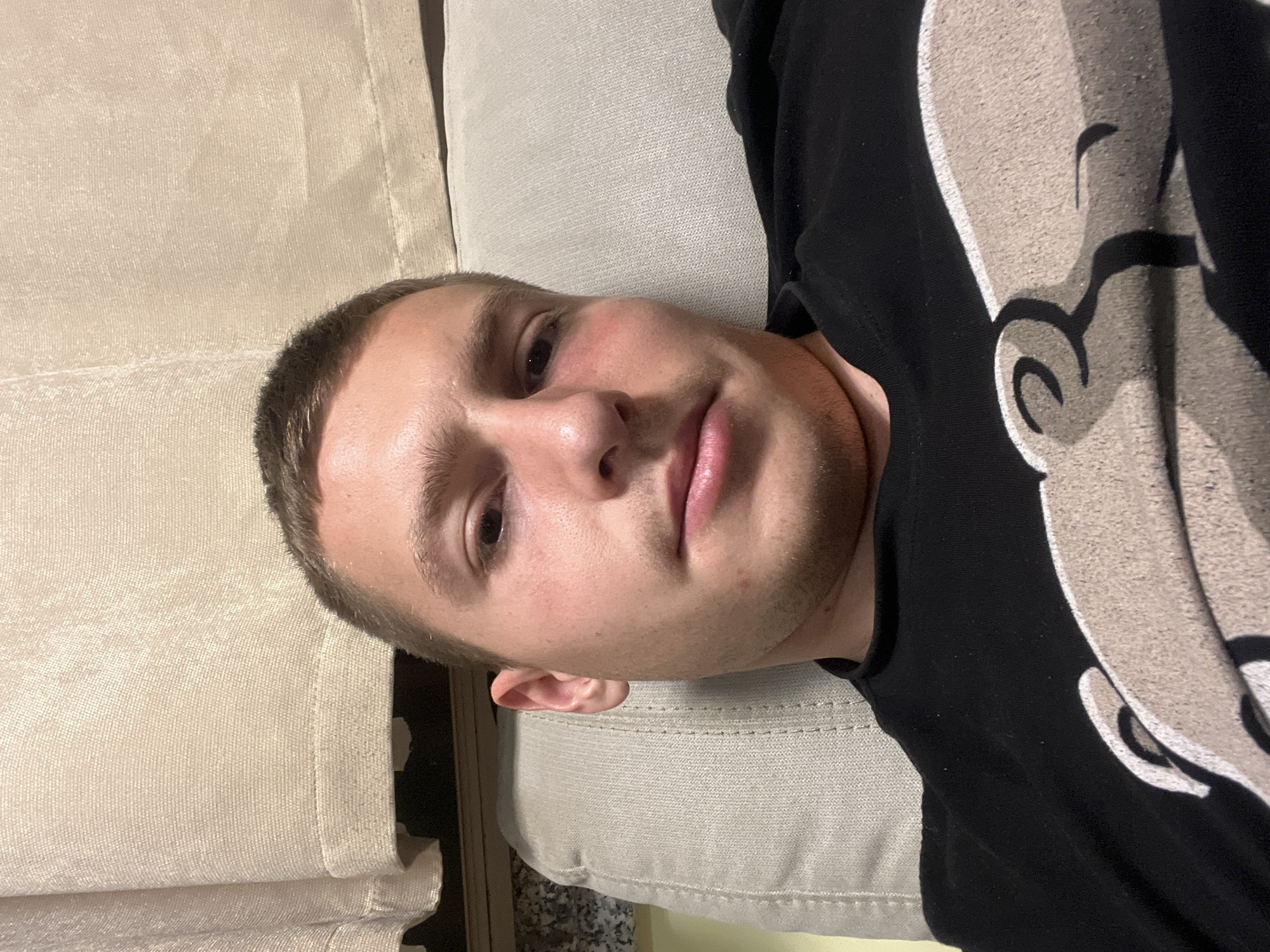 A light-skinned male with short brown hair is lying on a light beige textured surface, possibly a couch or bed. He is wearing a black shirt with a white graphic design on the right side of the image. The left side of the image shows a beige textured curtain-like material. The person appears to be looking directly at the camera. There is no indication of the location being Burgas, Bulgaria.