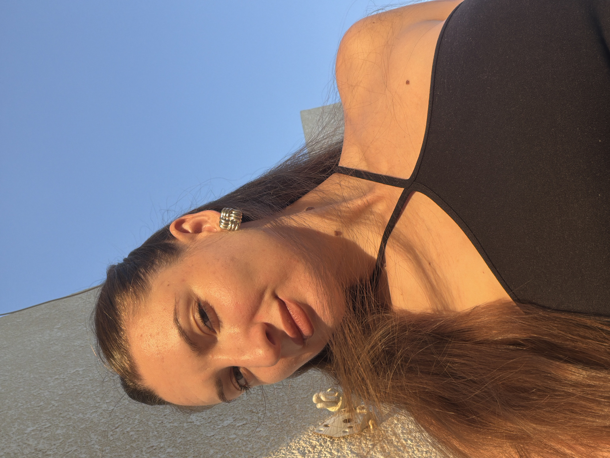 In Sharm El Sheikh, Egypt, a woman with long brown hair and brown eyes is captured in a close-up selfie against a clear blue sky. She is wearing a black tank top and a square, textured gold earring in her right ear. The woman is leaning against a textured, light-colored wall, which is illuminated by the warm glow of the sun. A decorative white element, possibly a light fixture or architectural detail, is visible on the wall to her left. Her skin is tanned, and her hair cascades over her shoulder and chest. The sun casts a warm, golden light across her face and upper body, highlighting her features and creating a soft focus on the background.