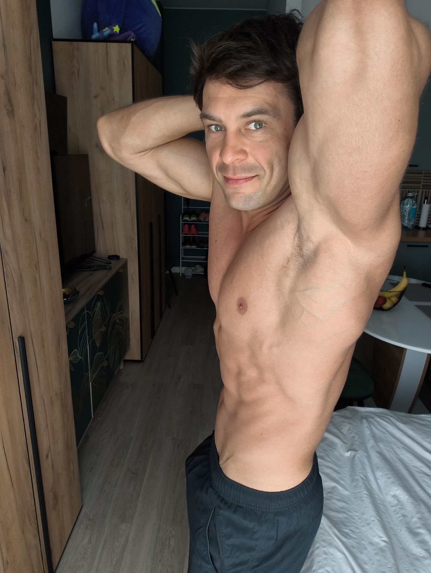 A muscular, shirtless man is posing for a photograph indoors. He is wearing dark athletic shorts and has his hands behind his head, flexing his biceps. The background includes wooden cabinets, a table with fruit, a bed with white sheets, and a glimpse of a hallway with shoes on a rack. The location is not discernible as being in Moscow, Russia without further context.
