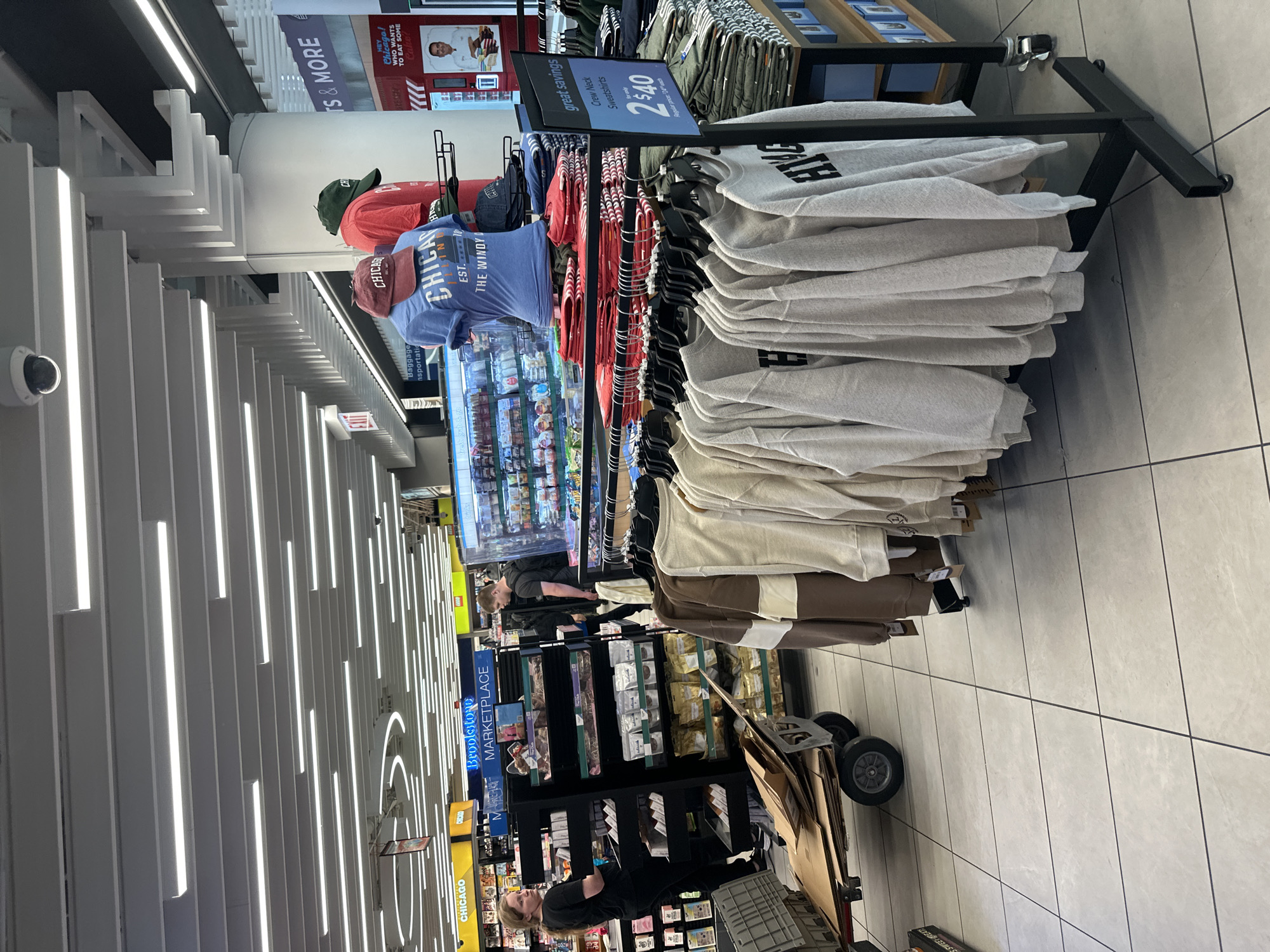 In Chicago, Illinois, USA, a retail store interior is depicted, focusing on a merchandise display. In the foreground, several grey crewneck sweatshirts are hung on a black metal rack, some bearing the text "HYE" in black. To the right, a display rack holds stacked green t-shirts. A sign on the sweatshirt rack advertises "great savings" on "Crew Neck Sweatshirts" for "2 $40". Behind this, mannequins display Chicago-themed apparel, including a blue t-shirt with "CHICAGO" and "EST. THE WINDY CITY" printed on it, and a red t-shirt with a green baseball cap. Further in the background, shelves are stocked with various products, including what appears to be food items, and signage indicating "Brookstone MARKETPLACE" and "CHICAGO". A woman in a black shirt and pants is visible on the left, pushing a wheeled cart loaded with cardboard boxes. A man with a backpack is standing near the shelves in the mid-ground. The environment features a tiled floor, a modern suspended ceiling with linear lighting, and a security camera mounted on the ceiling.