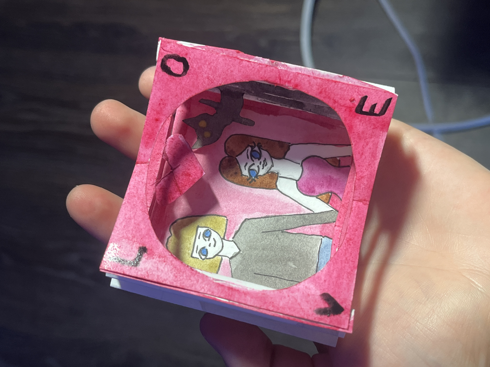 A small, hand-crafted diorama is held in the palm of a fair-skinned hand. The diorama is square, made of paper, and painted pink. A circular opening in the top layer reveals an interior scene depicted in a cartoonish style. The scene includes two female figures with blue eyes, one with blonde hair and one with brown hair. The blonde figure is wearing a grey top and blue jeans, while the brown-haired figure is in a pink dress. A black cat with yellow eyes is depicted in the background. Black symbols are painted on the exterior of the diorama, including what appears to be an "O", a "W", and an inverted "L" shape. The background is a dark, textured surface, and a light blue cable is visible in the upper right corner. The diorama appears to be a homemade craft project. There is no indication of Saint Petersburg, Russia, in the provided image.