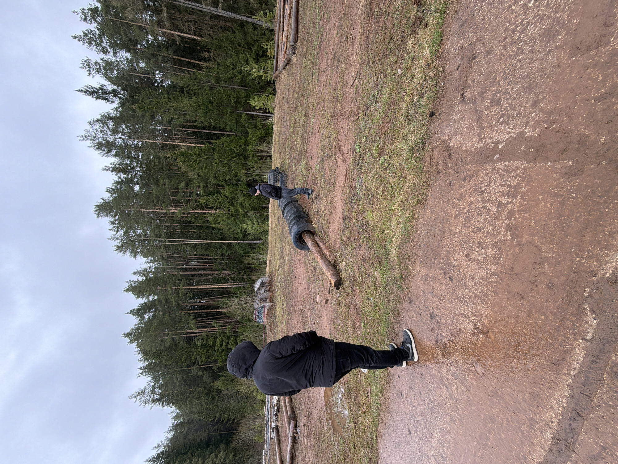 The image shows an outdoor scene in Прикамский, Russia. The environment consists of a muddy dirt path adjacent to a grassy area. A dense forest stands in the background. There are logs and tires strewn across the scene. Two individuals are visible: one is standing on the ground and wearing a black hooded jacket and black pants with white-soled shoes. The other person is standing on tires further away. The sky is overcast.