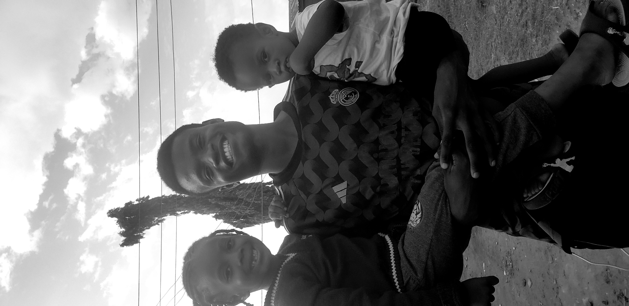 In Narayi, Nigeria, a young Black man stands outdoors, holding two young Black children. The man is in the center, smiling broadly at the camera, and is dressed in a patterned t-shirt. He is holding a small child, also a Black boy, on his right side, cradling him with his arms. The boy is wearing a t-shirt and shorts, and is looking at the camera with a slightly serious expression, with his hand near his mouth. On the man's left side, a young Black girl is held. She is wearing a sweater and pants, and is also looking at the camera, smiling. The background shows a tree and utility lines against a cloudy sky. The ground appears to be dirt.