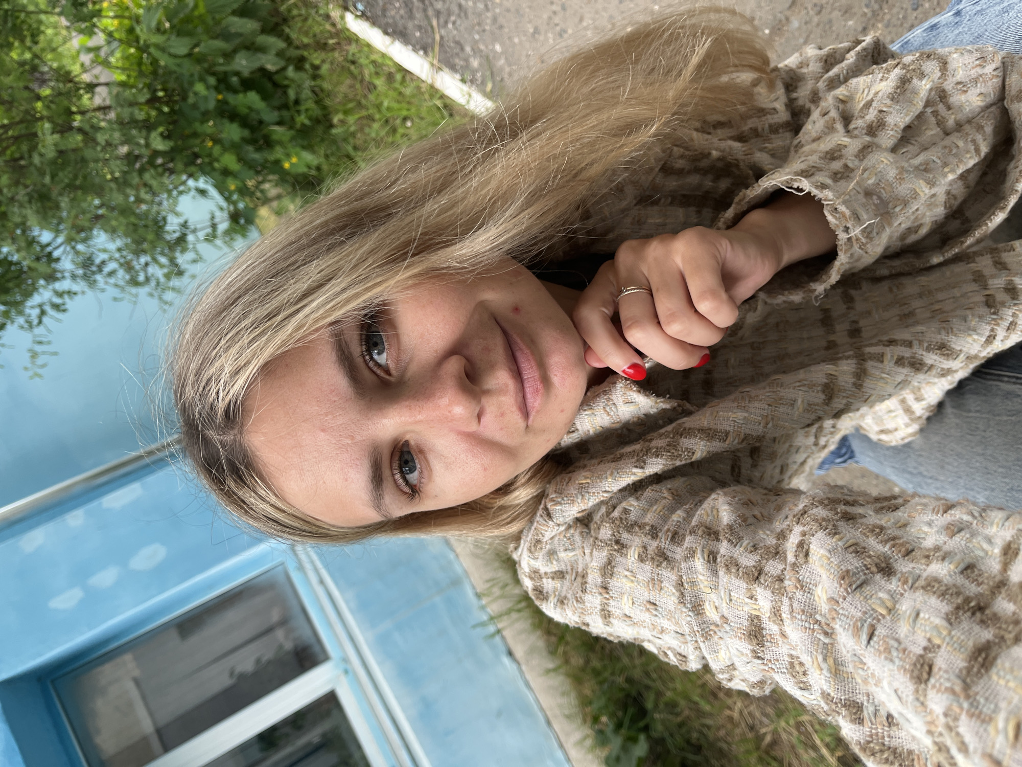 A candid, overhead, close-up selfie of a young Caucasian woman with long blonde hair and blue eyes, taken outdoors. She is wearing a textured beige and brown tweed jacket and blue jeans. Her left hand, adorned with red nail polish and a thin ring, is resting on her chin. The background features a bright blue wall with a window on the left, and green foliage and a grey paved surface on the right. The scene appears to be in the city of Chaikovsky, Russia, with the architecture and greenery characteristic of the region.
