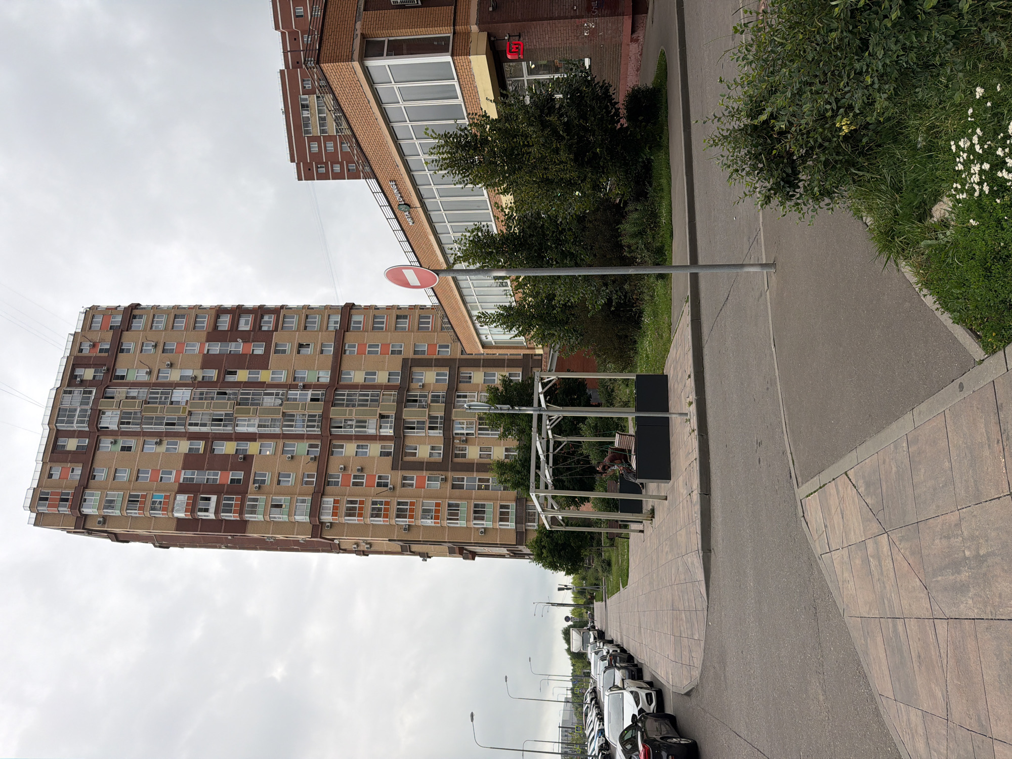 The image depicts an urban scene in Shcherbinka, Russia, characterized by residential buildings, a street, and a sidewalk. Dominating the view is a tall, multi-story apartment building with a facade of brown and tan bricks, accented by colorful window frames in shades of orange, yellow, and green. To the right, a lower, brick building with a contemporary design features a row of large windows, and a sign above them reads "MIATA LOUNGE." Adjacent to this building is a red square sign with a white horizontal bar, indicating a prohibition, likely a "do not enter" or "no stopping" sign.
The street is lined with parked cars, primarily white and black sedans, suggesting a residential area. A paved sidewalk, laid with large square tiles, runs along the street. In front of the brick building, a small park-like area with trees and benches is visible. A person is seated on a bench within a modern, open-air structure with a metallic frame, appearing to be a small canopy with swings. The sky is overcast with gray clouds. The overall environment appears to be a relatively modern urban development.