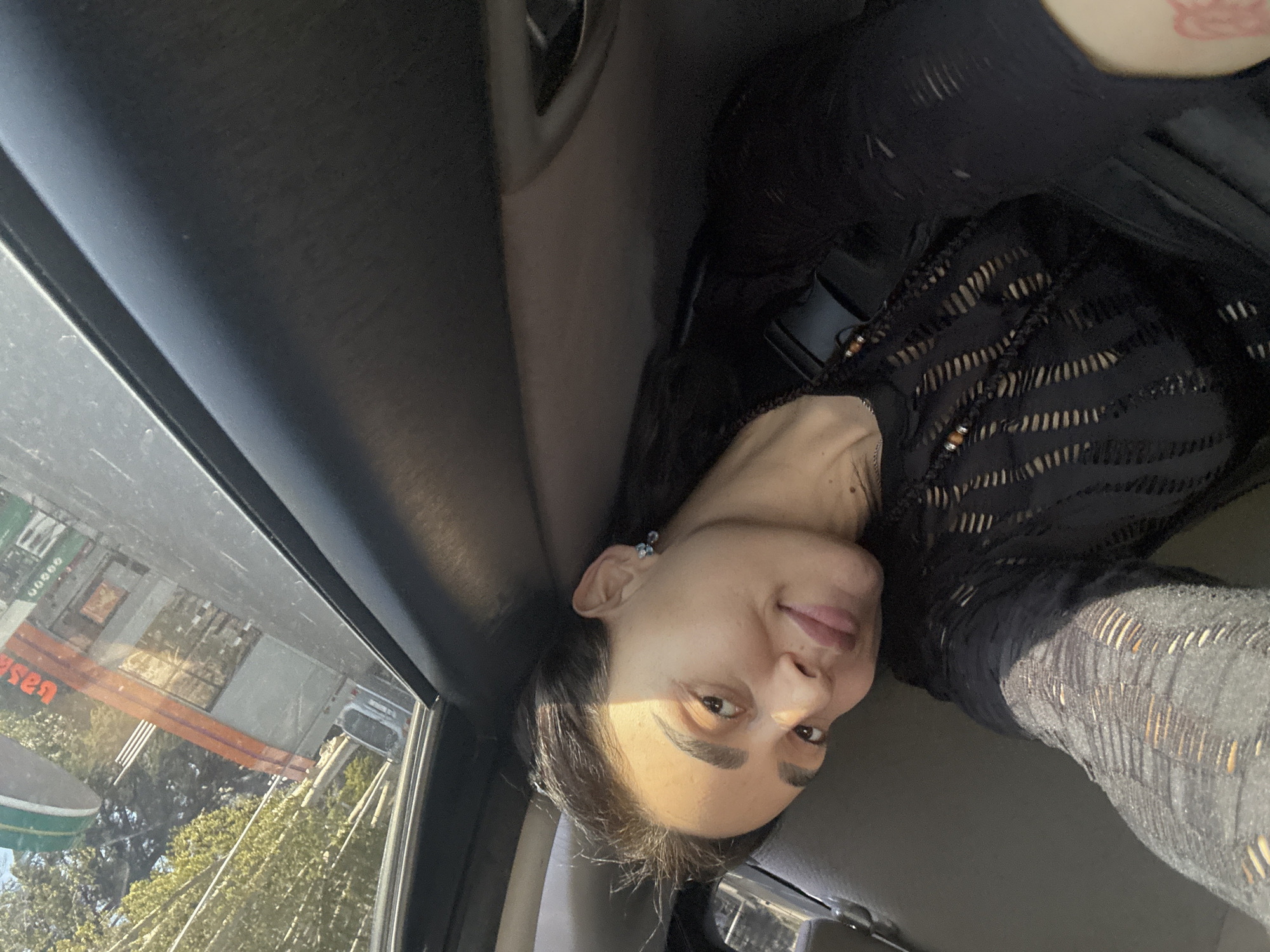 A woman is sitting inside a vehicle, likely a car, with the interior visible including parts of the dashboard, door, and window frame. The exterior, visible through the window, suggests an urban environment. There are buildings, trees, and what appears to be a street sign. Based on the sign, it seems the location is the city of Tbilisi, Georgia. The woman is wearing a dark-colored, possibly lace-patterned or striped garment. She is turned towards the camera, offering a partial view of her face. The lighting suggests it's daytime.