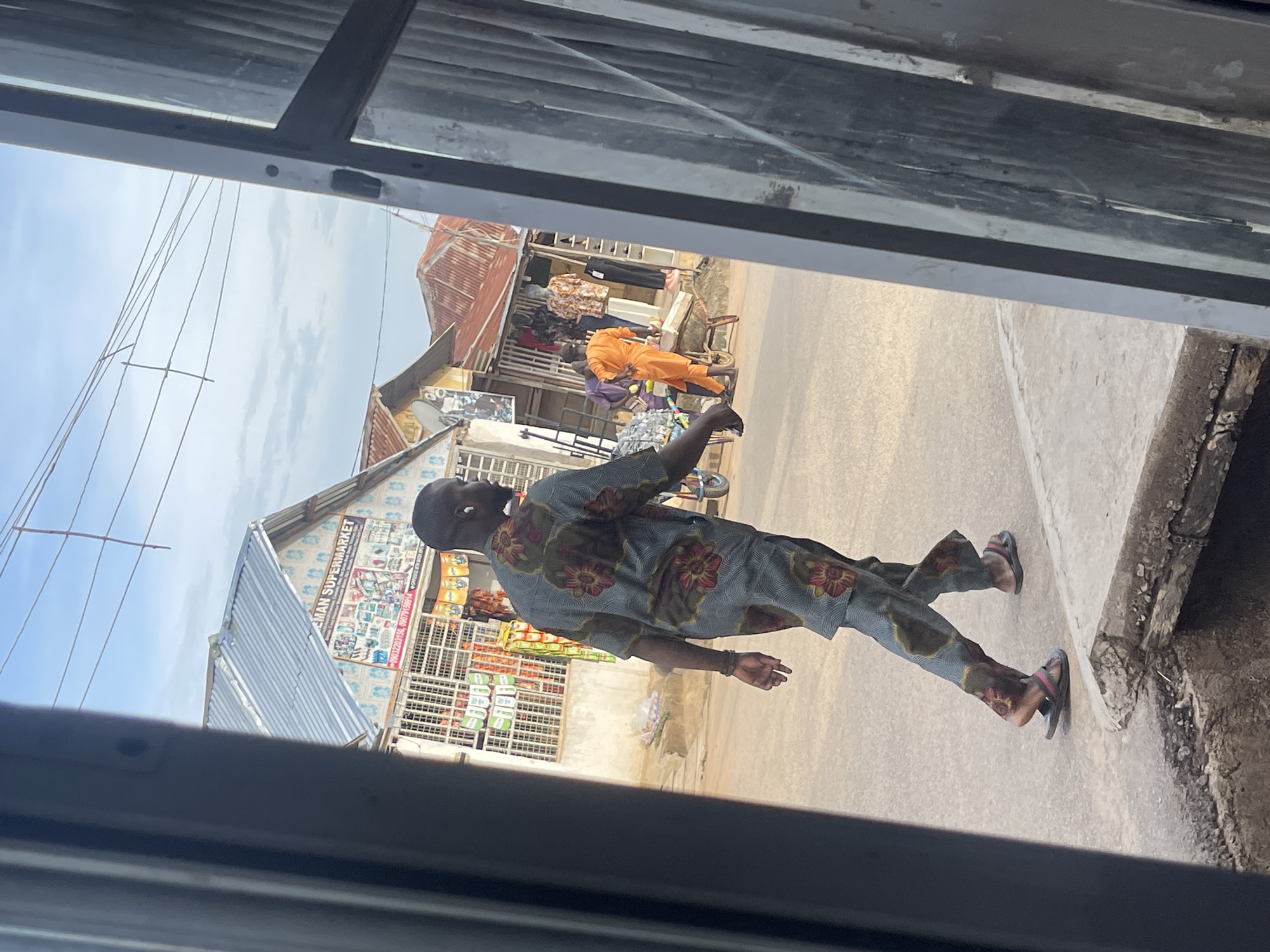 This photograph, taken in Abuja, Nigeria, presents a street scene viewed through a partial frame, with dark vertical elements visible on the far left and right. In the foreground, a man with short dark hair and a beard walks away from the camera, slightly right of center. He is dressed in a two-piece patterned outfit featuring gray-blue fabric with red and olive-green floral or abstract designs. A white wireless earbud is visible in his right ear. He wears dark flip-flops, with a pink strap visible on his left foot. He is walking on a paved surface next to a concrete curb, below which a small broken section or drain opening is visible.
On the left side of the street, a single-story building with a light gray corrugated metal roof displays a prominent sign reading "AMAN SUPERMARKET." Below the supermarket name, a collage of product images (including footwear and bags) is visible, along with two phone numbers: "09032203150" and "09079139814." The lower part of this building features a grated facade, behind which various items are discernible. Further down the street, on the right, another man dressed in a bright orange two-piece garment stands near a wheelbarrow. Nearby, several commercial structures with rust-colored corrugated metal roofs house open-front shops, displaying clothing on hangers and mannequins, one of which wears a patterned shirt. A white satellite dish is mounted on the roof of one of these structures. Overhead, multiple black utility wires span across a light blue sky dotted with scattered white clouds.