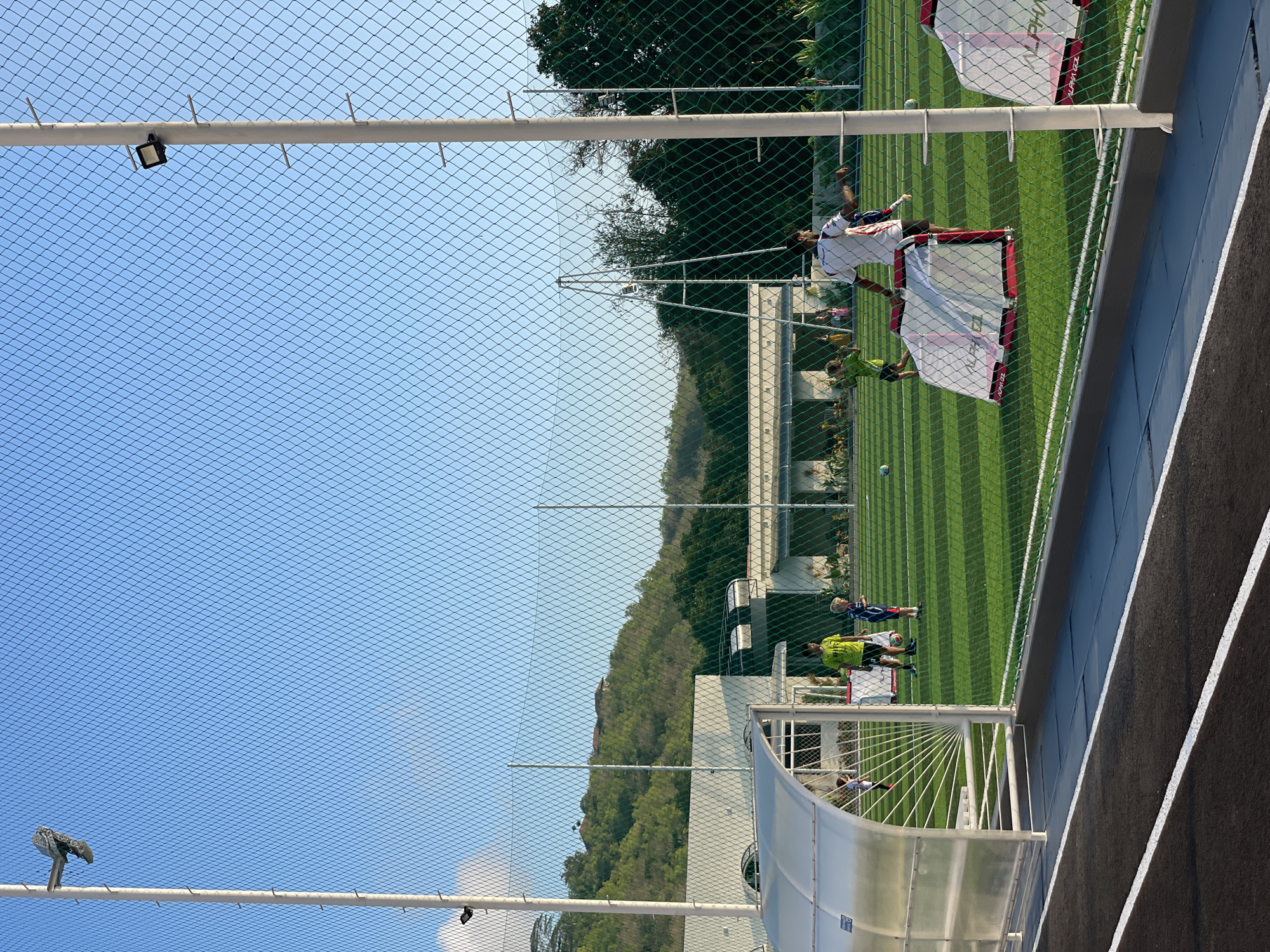 The image depicts an outdoor artificial turf soccer field under a clear blue sky, likely in Kuta Selatan, Indonesia, given the tropical vegetation visible on the hills in the background. The field is lined with white markings, and two small portable soccer goals with red trim are visible on the right side of the frame. A group of people, primarily children, are scattered across the field, some appearing to be engaged in a soccer practice or game. One adult, wearing a white and red shirt, stands near the goals. A covered bench or dugout with a translucent roof is situated on the left side of the frame, with at least one child visible underneath it. A chain-link fence runs along the foreground, separating the spectator area or track from the field. Light poles with attached spotlights are visible along the fence line, suggesting the field is equipped for night use. The overall scene conveys a sense of organized sporting activity in a well-maintained outdoor facility.