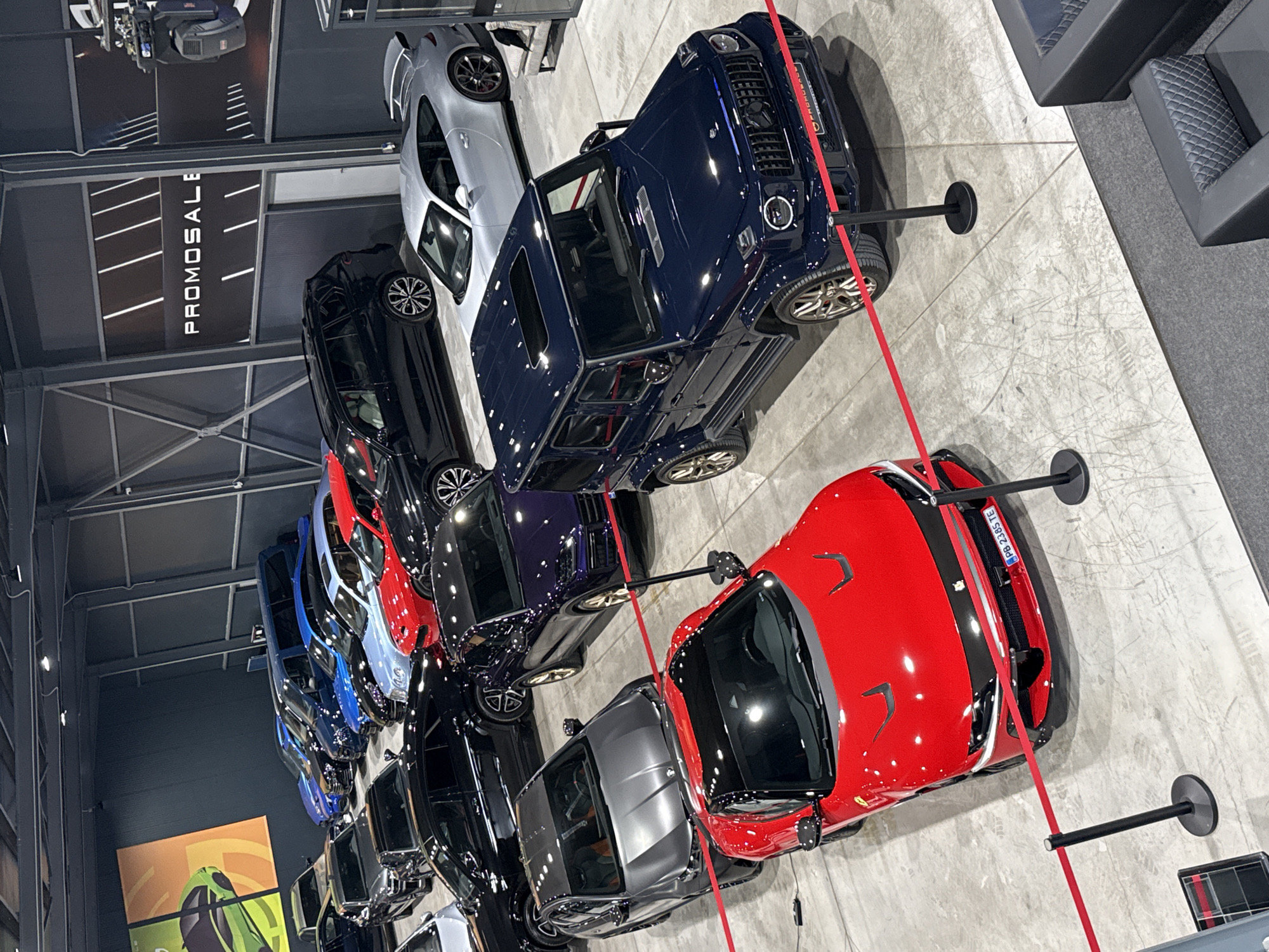 The image depicts a car dealership in Plovdiv, Bulgaria, showcasing a variety of luxury cars, including a red Ferrari, a dark blue Mercedes-Benz G-Class, and several other cars in shades of black, gray, and blue. The cars are parked on a light gray concrete floor and are sectioned off with red rope barriers held up by black stands. A sign that reads "PROMOSALE" hangs from the ceiling.