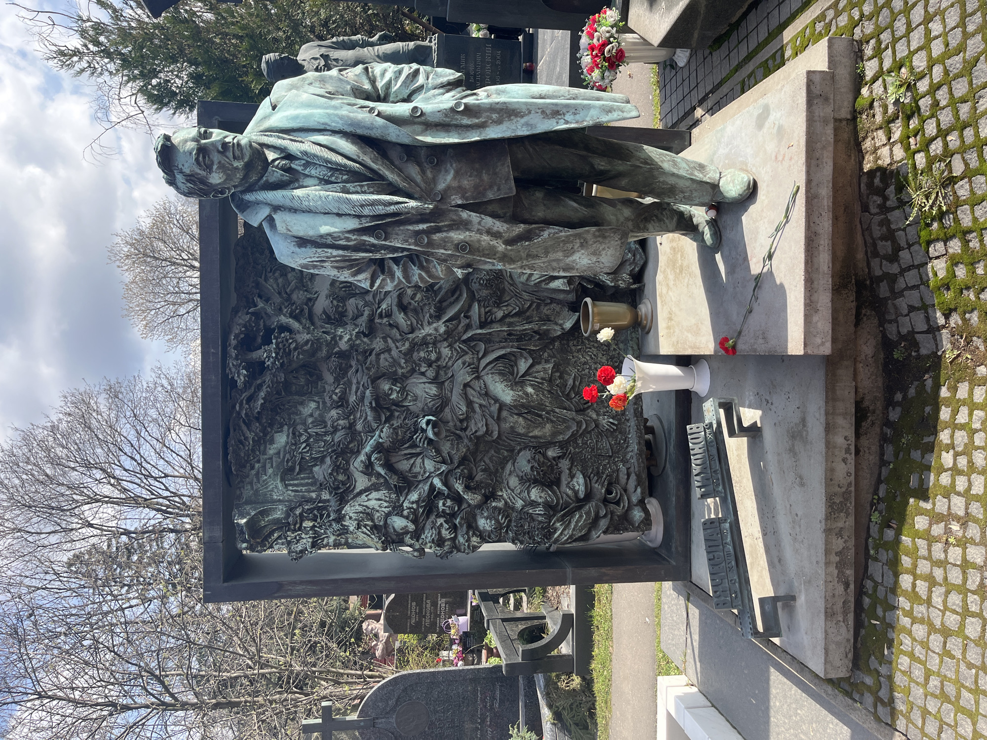 The image shows a monument at the Novodevichy Cemetery in Moscow, Russia. The monument depicts a bronze statue of a man in a coat standing in front of a bronze bas-relief panel. A vase with red and white flowers sits on the monument's base. The ground around the monument is paved with cobblestones and moss. Other tombstones and trees are visible in the background.