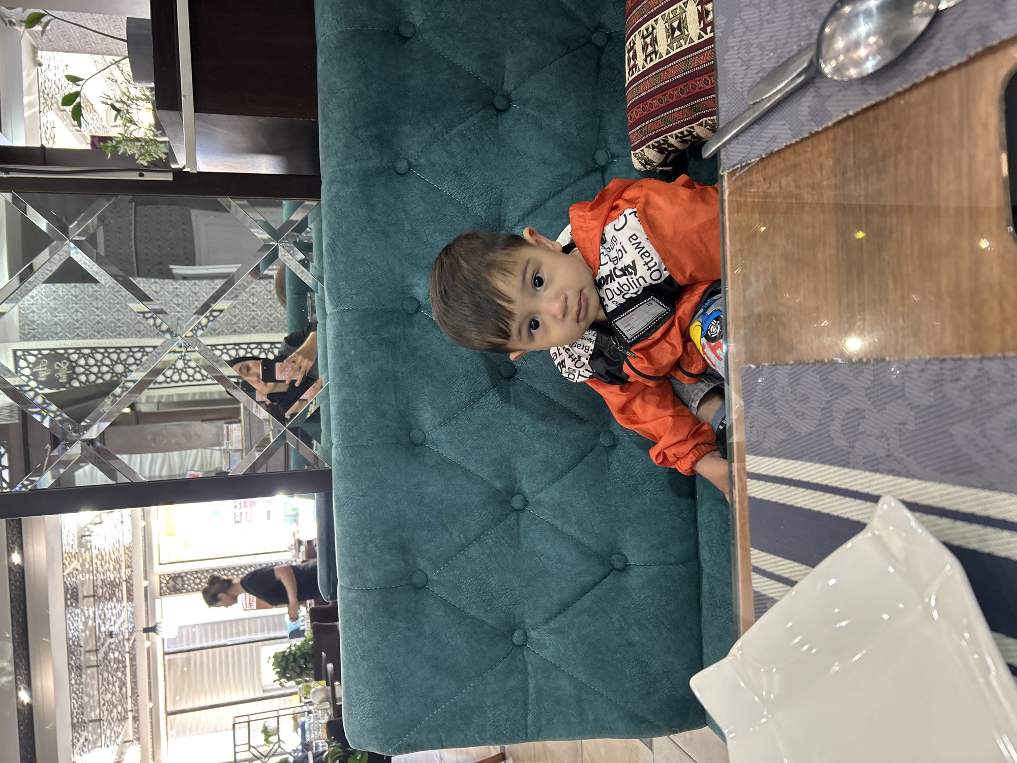 A young child wearing an orange jacket with white writing is seated on a teal tufted chair. There's a spoon and knife on a table with a grey patterned placemat. A white napkin is also on the table. The child is looking at the camera. Reflections in a decorative mirror show other people and the interior of a restaurant. The image was taken in Ақсу, Kazakhstan.