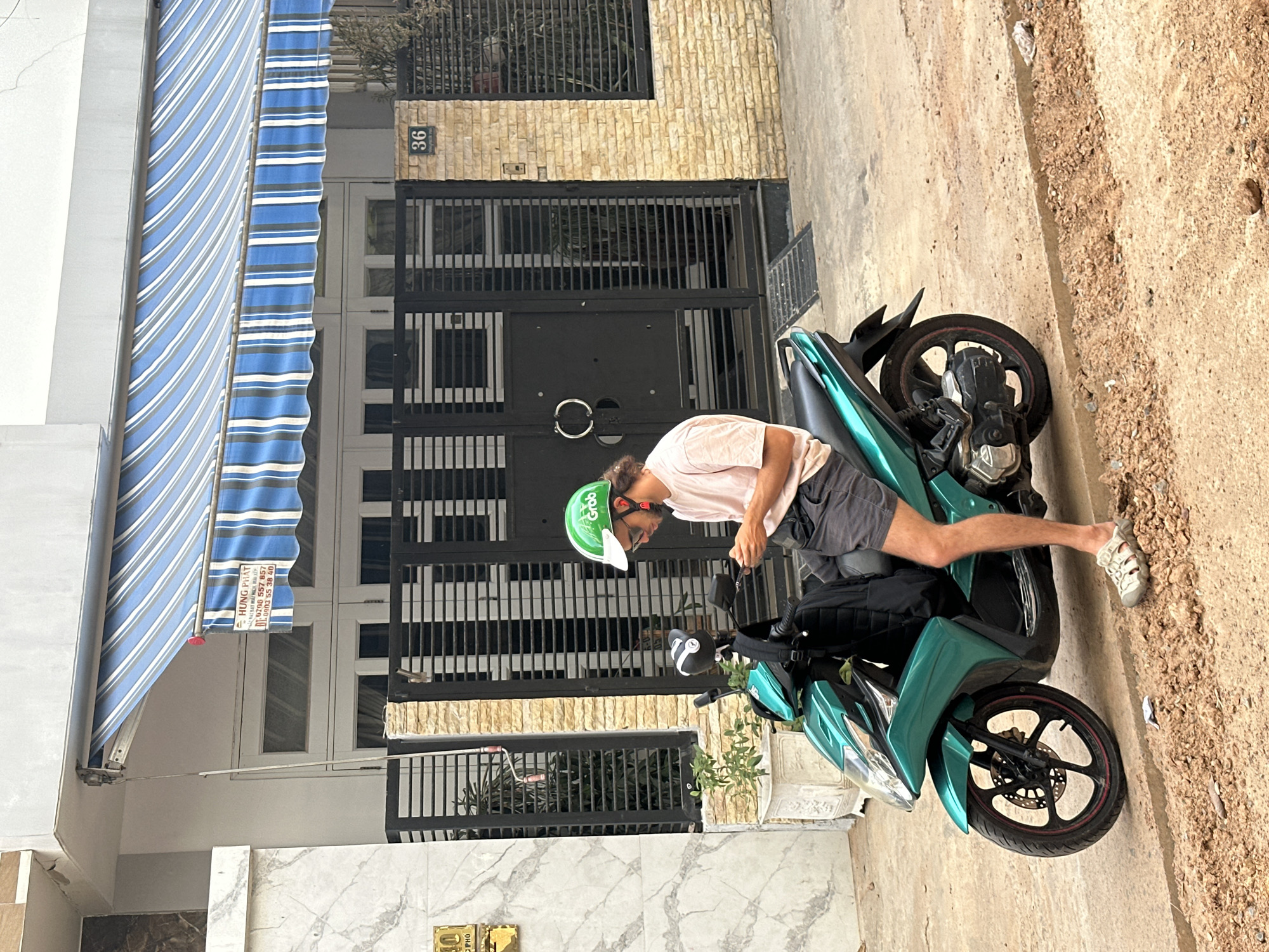 A person wearing a pink t-shirt, dark shorts, and a green Grab helmet is sitting on a teal motorcycle. They are holding what appears to be a phone or a small electronic device in their hands. The motorcycle is parked on a dirt and concrete path. Behind the motorcycle is a building with a black metal gate and a tan brick facade. The number "36" is visible above the gate. To the left of the building is a white section with a blue and white striped awning. The scene appears to be outdoors in a residential or commercial area. The lighting suggests it is daytime, possibly late morning or early afternoon.