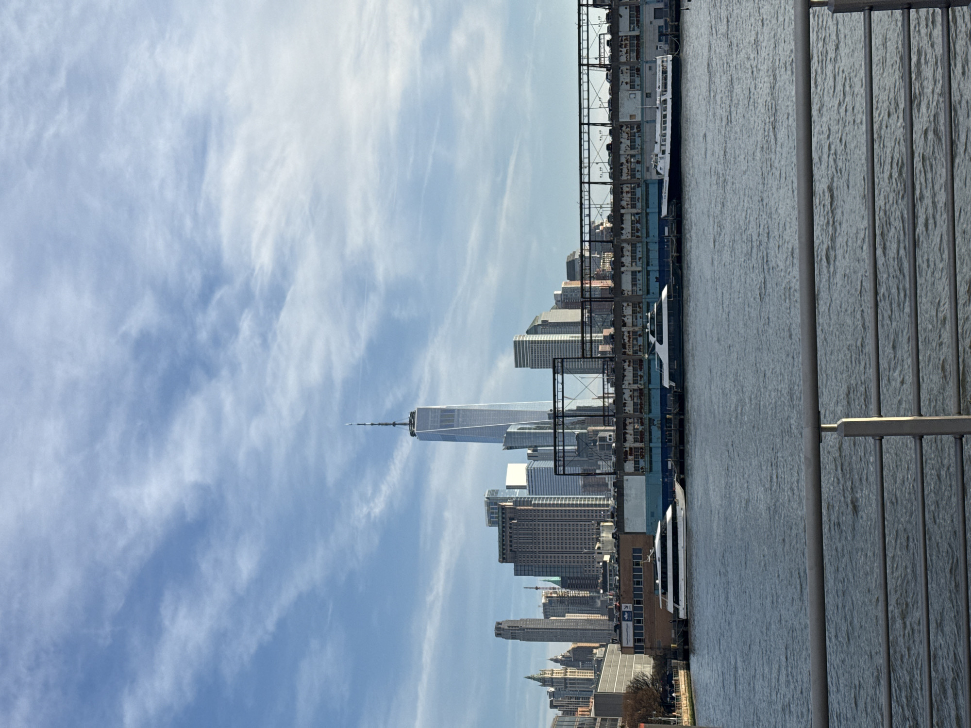 The image shows the New York City skyline under a blue sky with wispy clouds. The One World Trade Center is prominently visible amidst other skyscrapers. A body of water, possibly the East River or New York Bay, stretches across the foreground with docks and several boats docked along the edge. A fence with vertical bars is visible on the right side of the image, suggesting the viewpoint is from a pier or similar structure.